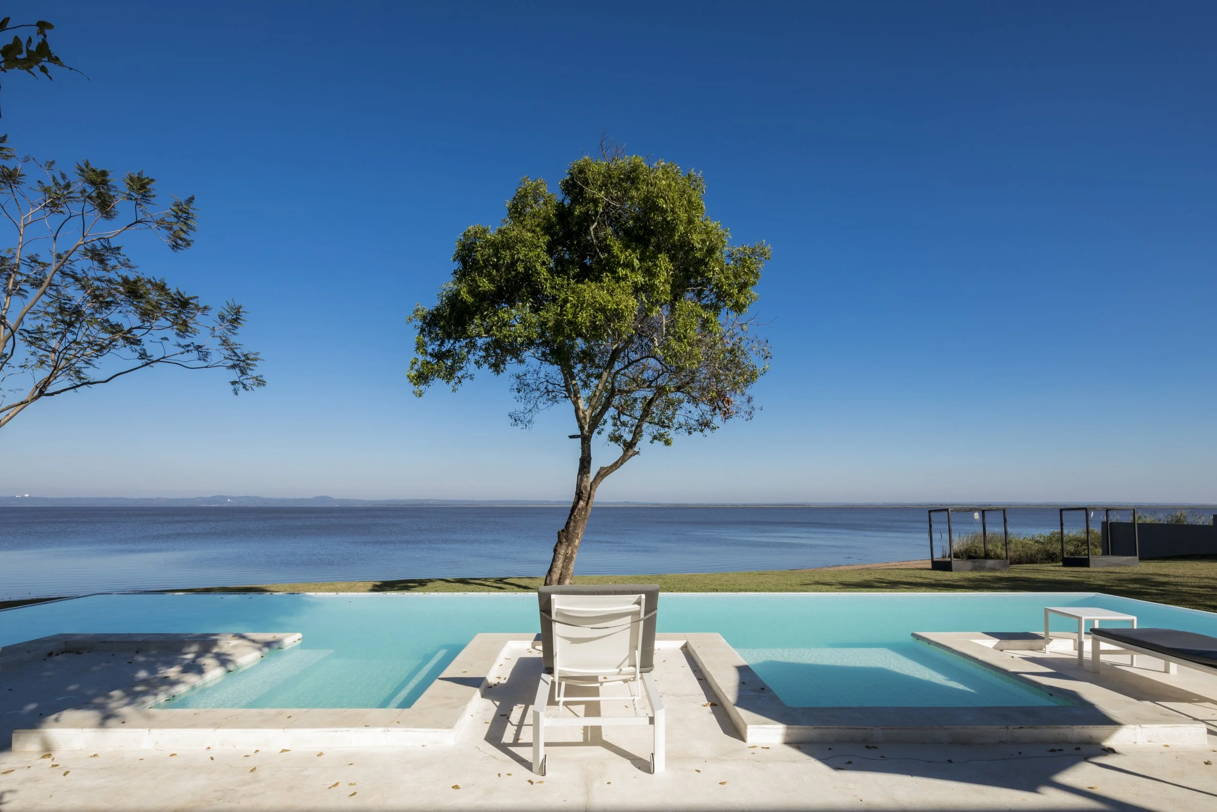 	
PRO ARQUITECTURA PARAGUAY architecture design PROSA
Vista de una piscina con una silla y un árbol al fondo, frente a un lago o mar bajo un cielo despejado.