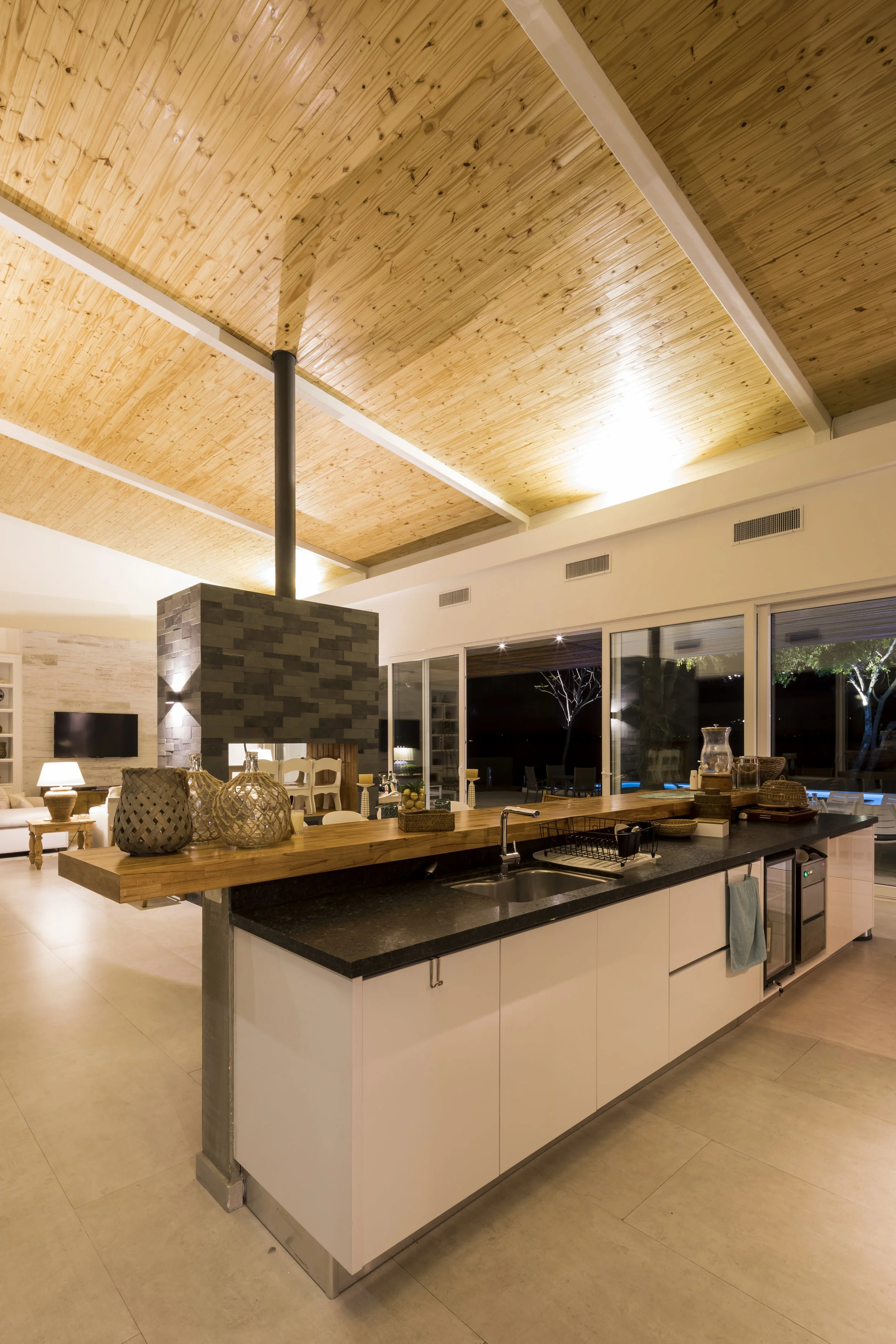 	
PRO ARQUITECTURA PARAGUAY architecture design PROSA
Cocina moderna en interior con encimera negra, lavabo y utensilios, techos de madera, área social visible con sillones y chimenea.