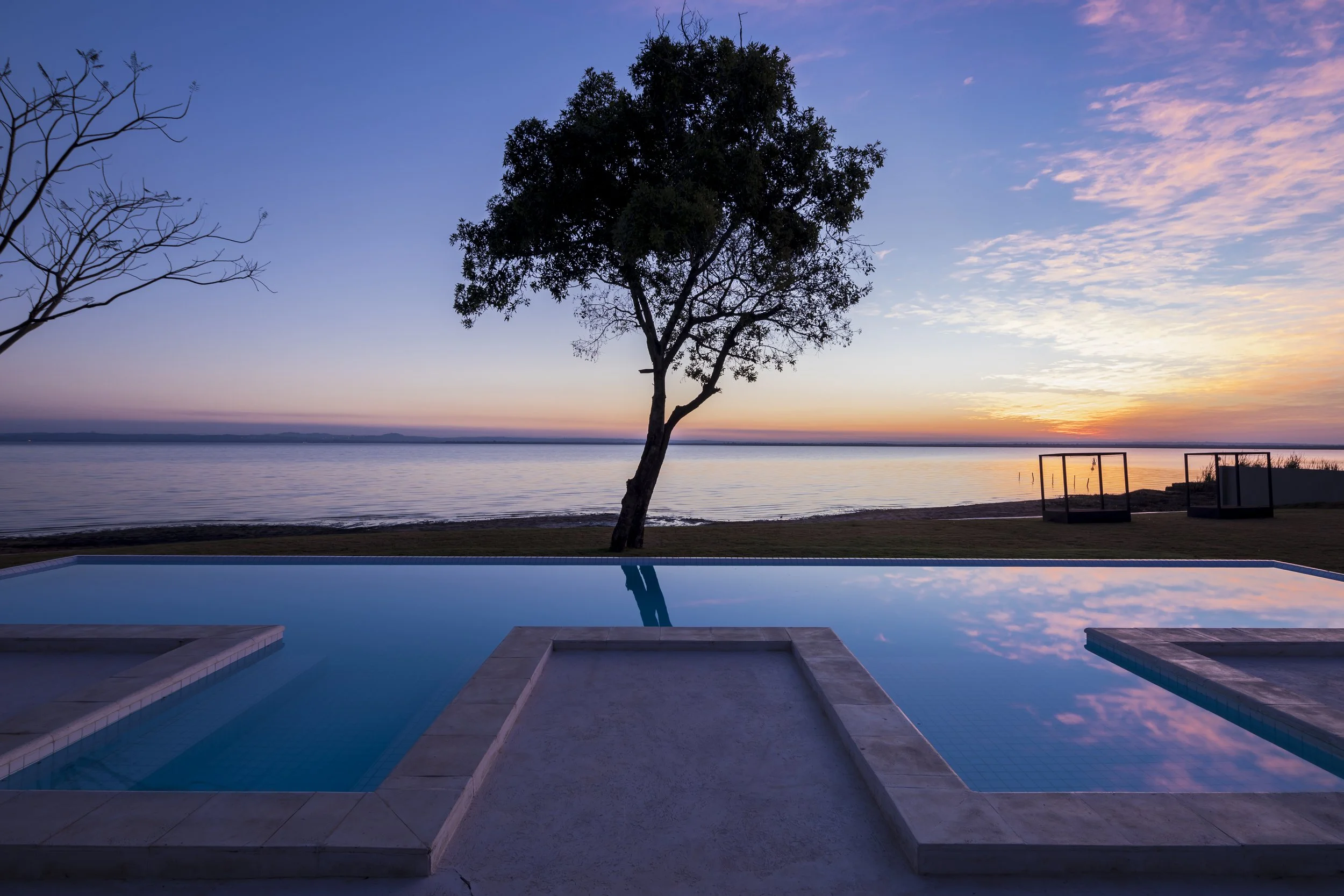 	
PRO ARQUITECTURA PARAGUAY architecture design PROSA
Piscina con vista al lago, árbol solitario y puesta de sol, con estanterías de madera en el fondo