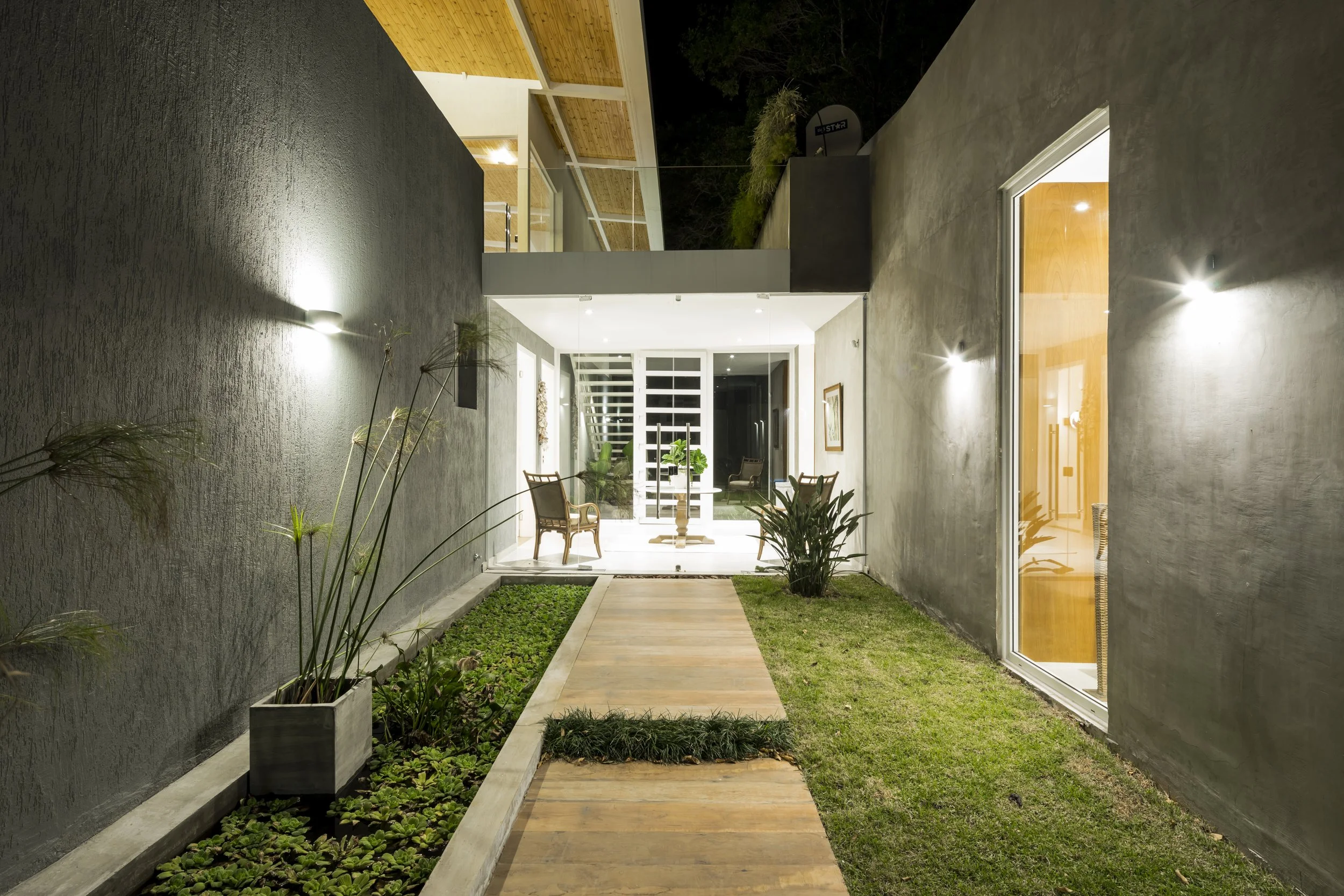 	
PRO ARQUITECTURA PARAGUAY architecture design PROSA
Pasillo exterior delgado con paredes grises, césped y plantas en macetas, iluminado por luces de pared, que conduce a una sala de interiores con muebles y decoraciones