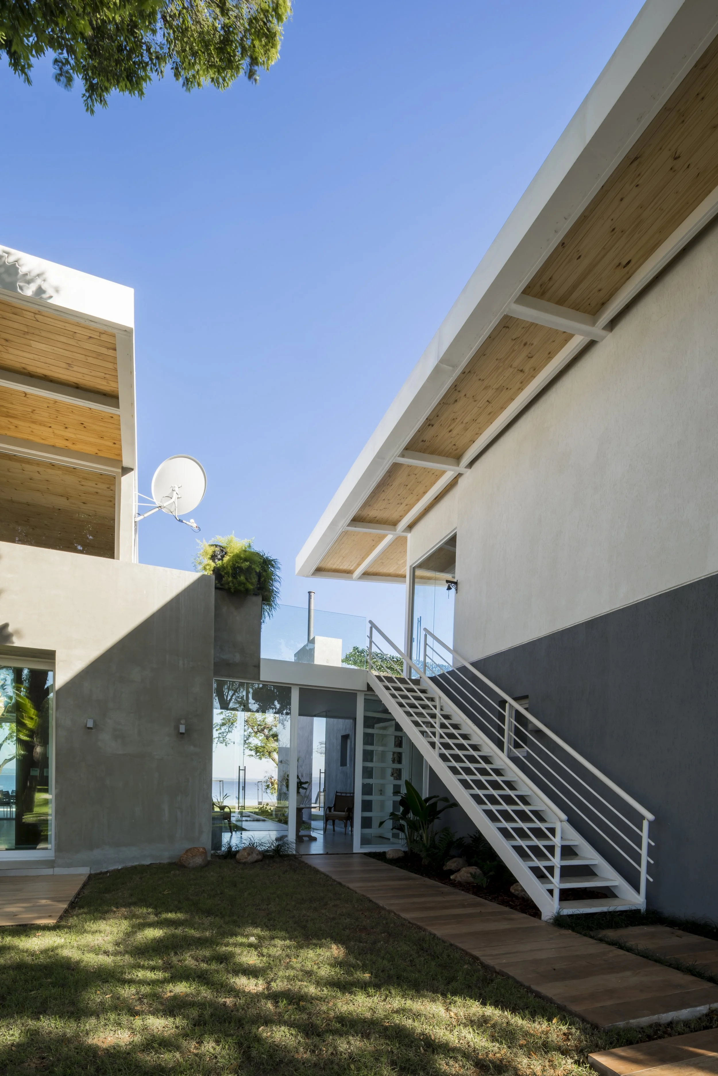 	
PRO ARQUITECTURA PARAGUAY architecture design PROSA
Casa moderna de dos pisos con escaleras exteriores, paredes de color gris claro y techo de madera, área de césped con camino de madera y plantas, en un día soleado con cielo azul.