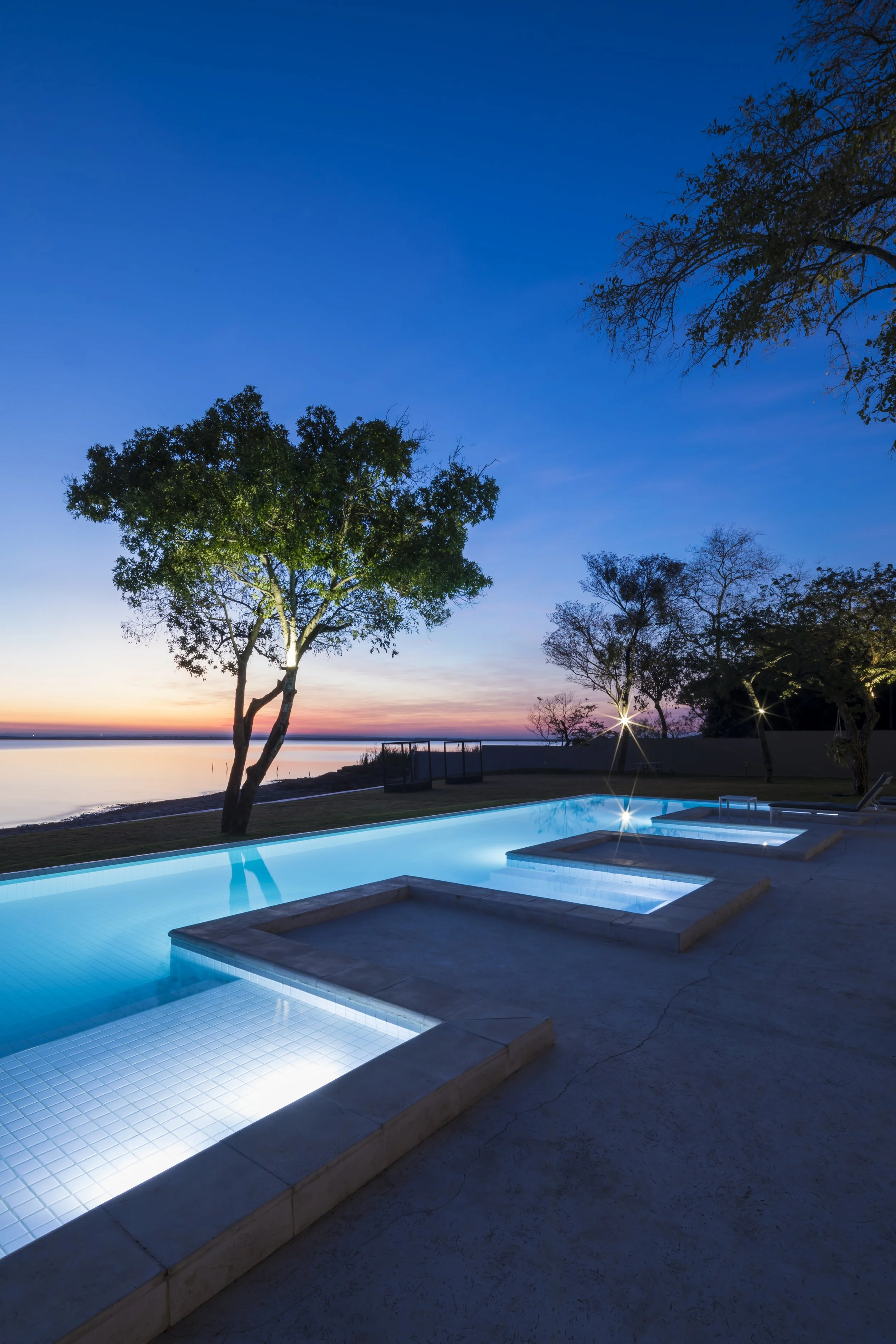 	
PRO ARQUITECTURA PARAGUAY architecture design PROSA
Piscina iluminada con luces en un entorno natural al atardecer, con árboles y un río o lago al fondo.