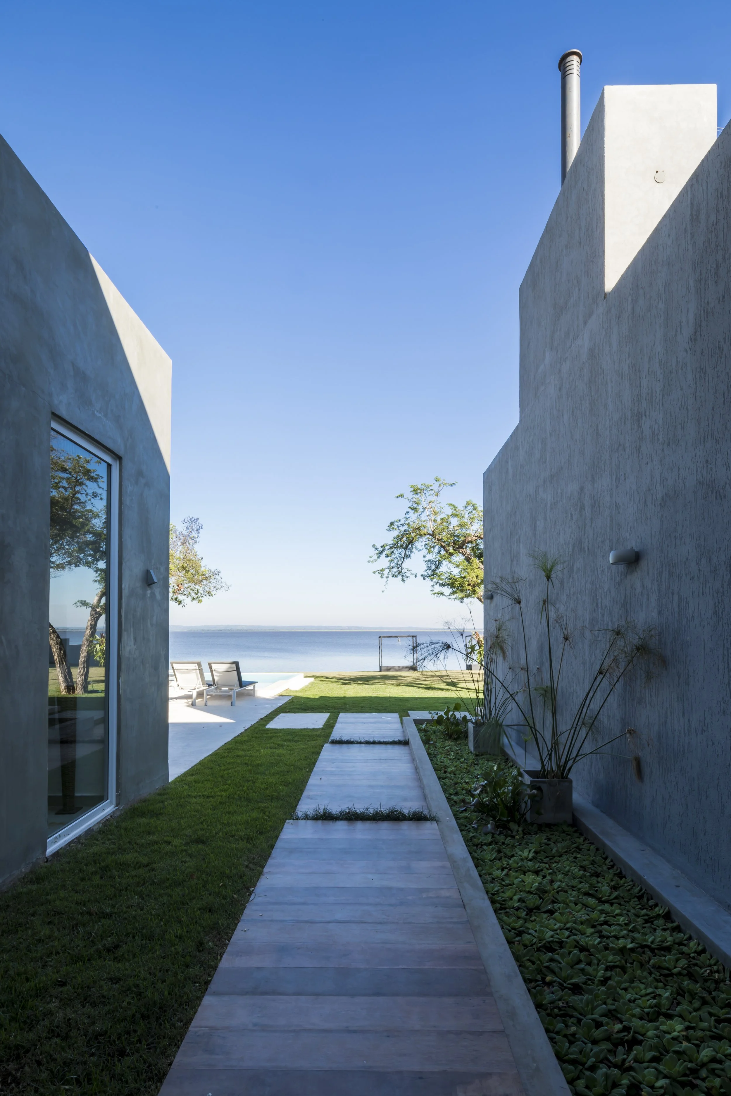Camino de piedra entre dos casas modernas con vista al mar y área de descanso con sillas junto a árboles y césped