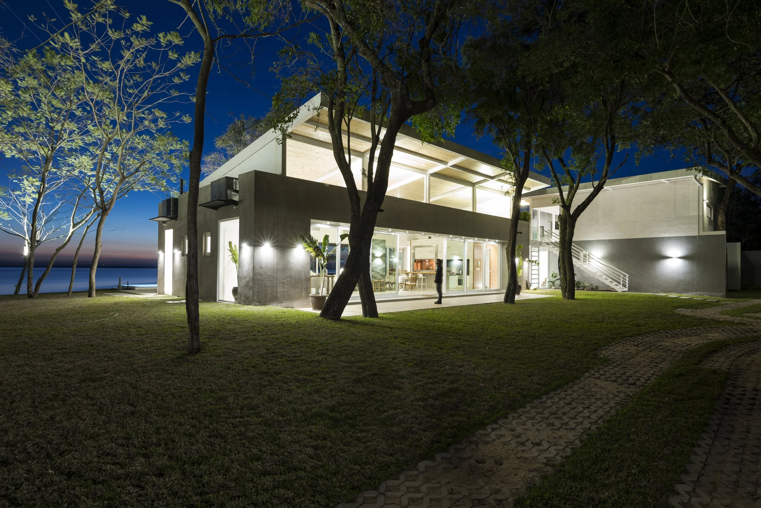 	
PRO ARQUITECTURA PARAGUAY architecture design PROSA
Casa moderna iluminada en la noche, rodeada de árboles y con vista al mar en el fondo.
