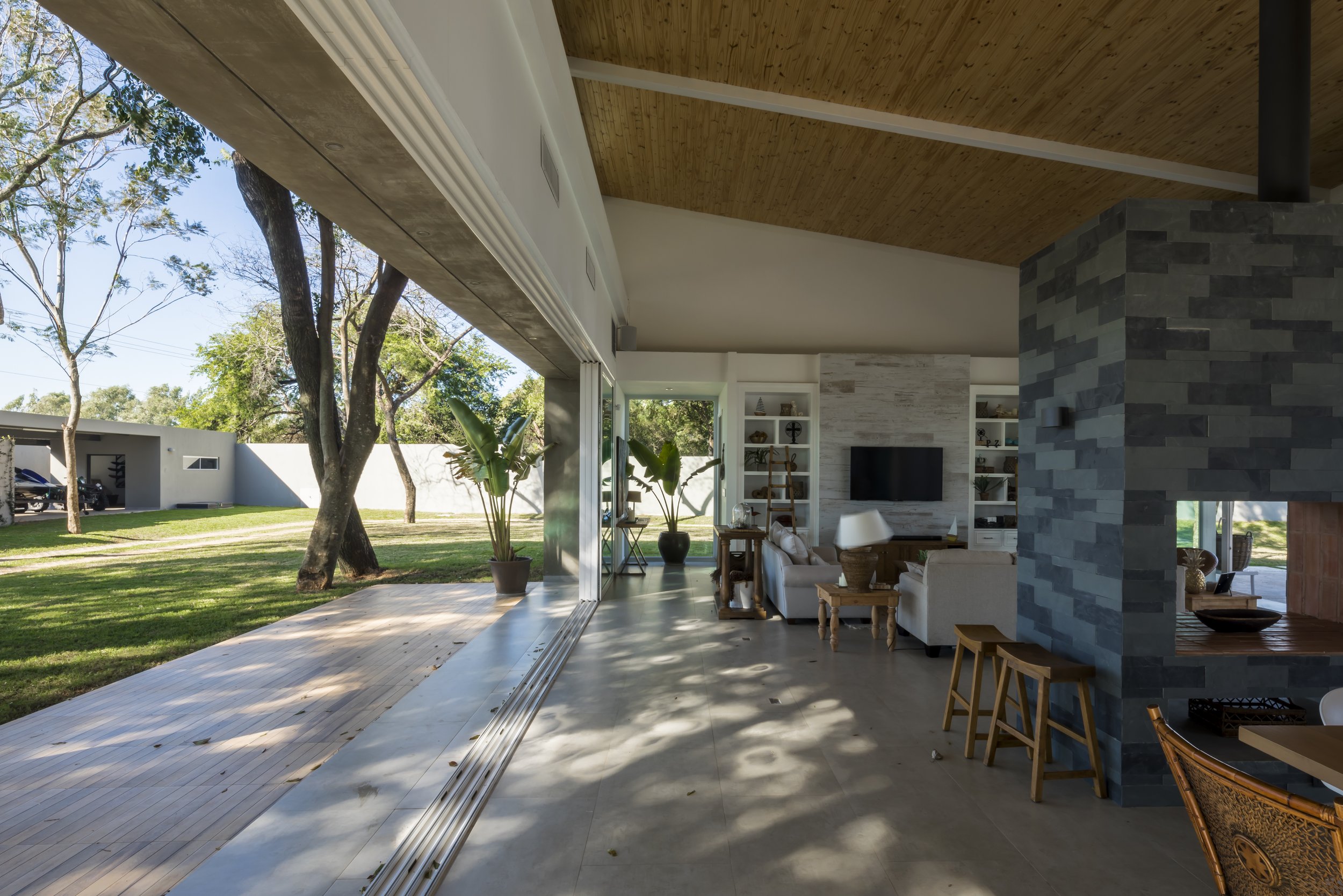 Interiores de una sala moderna con paredes de piedra azul, sillones blancos, estanterías con objetos decorativos y un televisor, con vista a un jardín con césped y árboles.