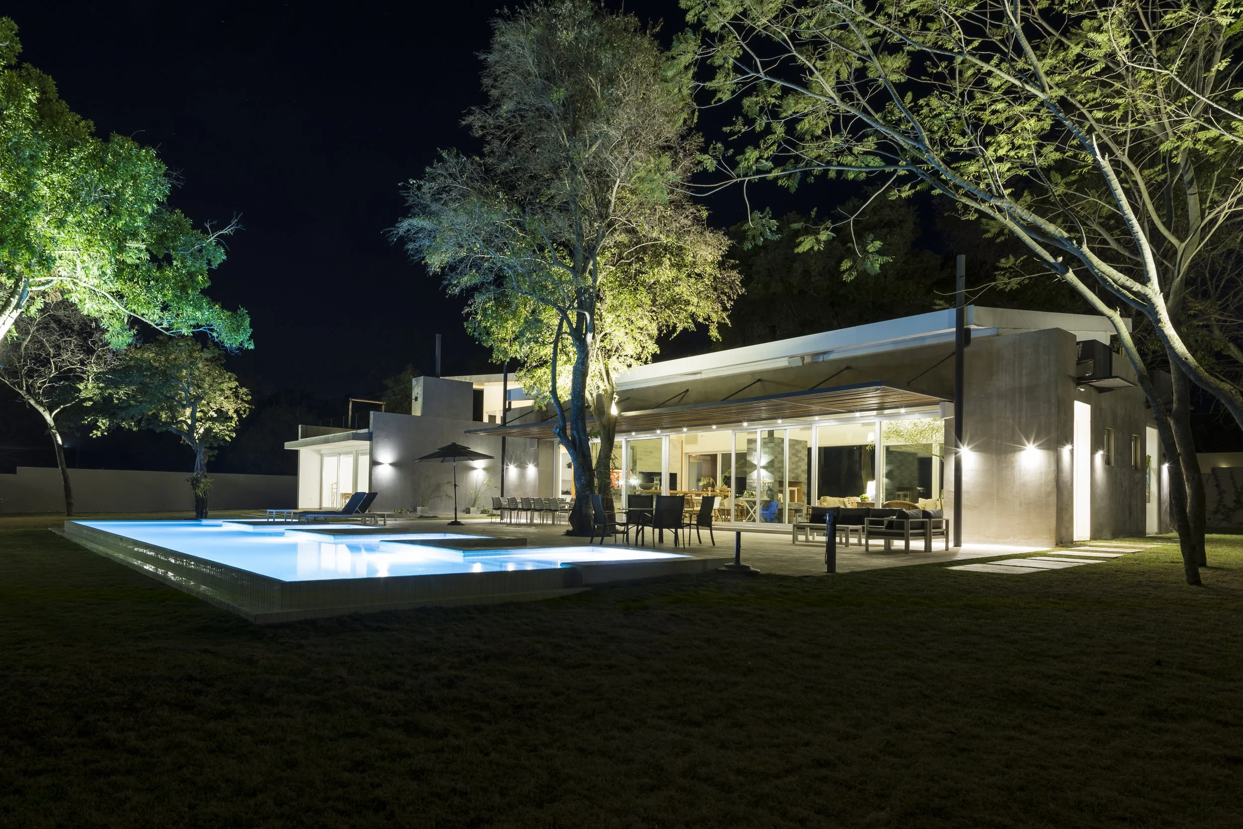 	
PRO ARQUITECTURA PARAGUAY architecture design PROSA
Casa moderna con iluminación exterior, piscina iluminada y árboles en el jardín en la noche.