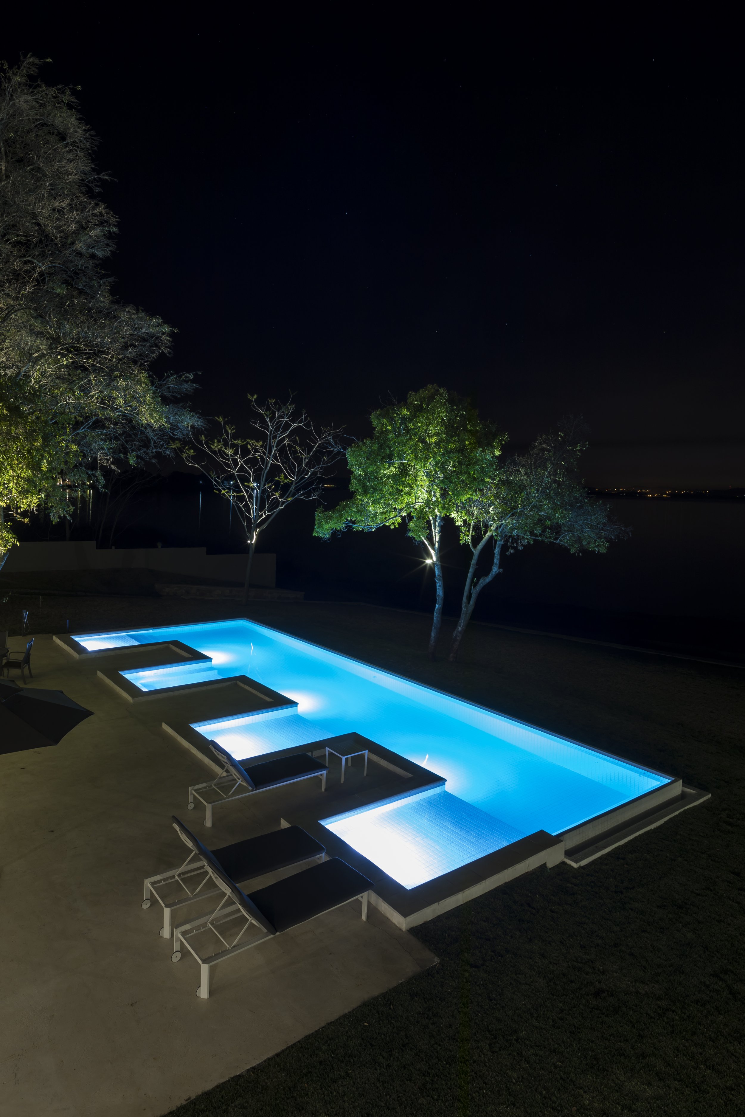 	
PRO ARQUITECTURA PARAGUAY architecture design PROSA
Piscina iluminada de noche con sillas y mesa, árboles y césped en el fondo.