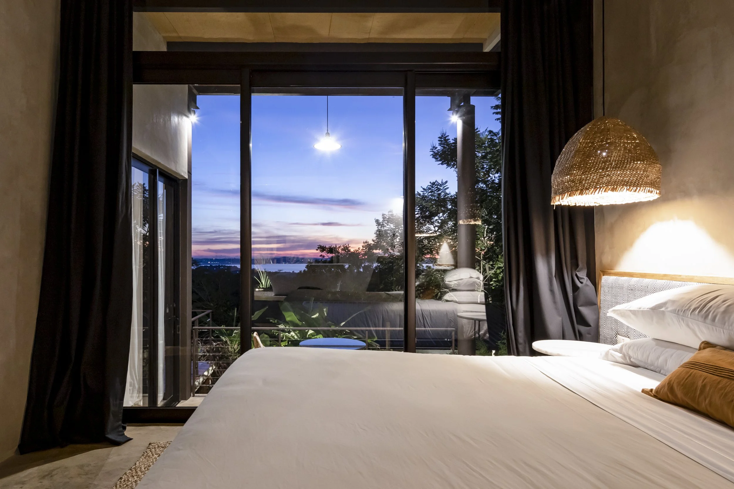Habitación con vista al atardecer, cama, cortinas negras, lámpara de techo de mimbre, ventanales grandes y balcón con plantas y sillón.