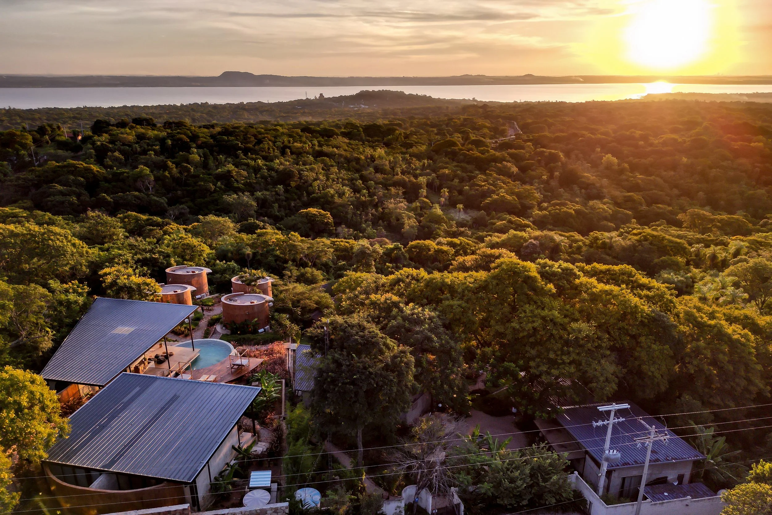 Vista aérea de casas con techos de metal entre árboles durante el atardecer, junto a un cuerpo de agua en el fondo.