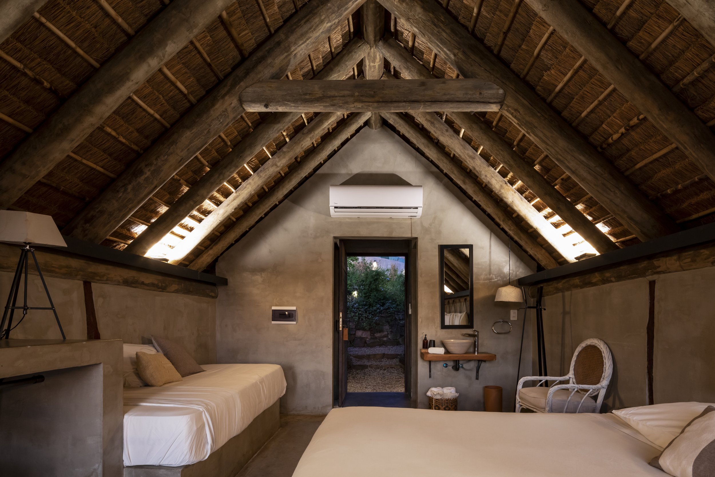 Dormitorio en una cabaña con paredes de cemento, techo de vigas de madera y tabiquería, con una cama doble y una cama individual, una lámpara de pie, un espejo, un ventilador de aire acondicionado y una silla junto a un lavabo y un espejo sobre una p