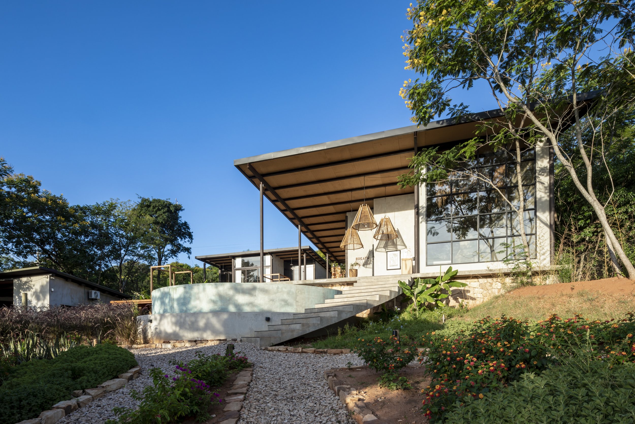 Casa moderna con grandes ventanas y techo inclinado, rodeada de jardín con plantas y árboles, en un día soleado