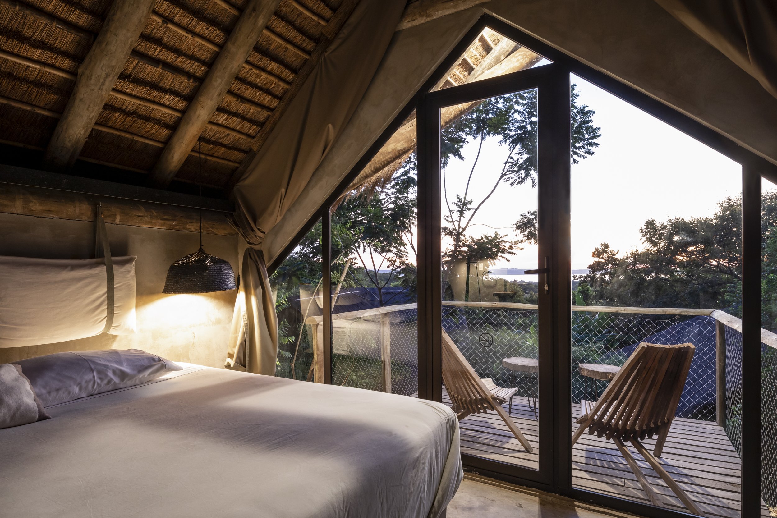 Habitación con cama, lámpara colgante, y balcón con asientos y vista a la naturaleza al atardecer.
