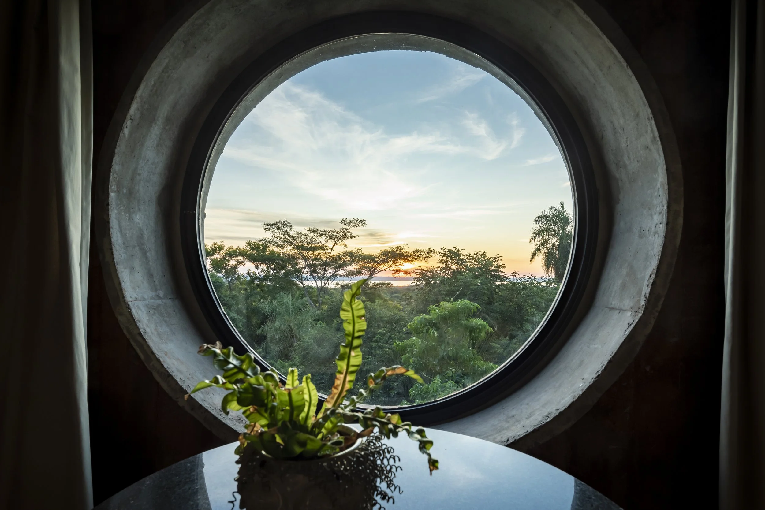 Vista de un atardecer desde una ventana redonda, con un florero con planta en primer plano. Se observa vegetación y el cielo con nubes.