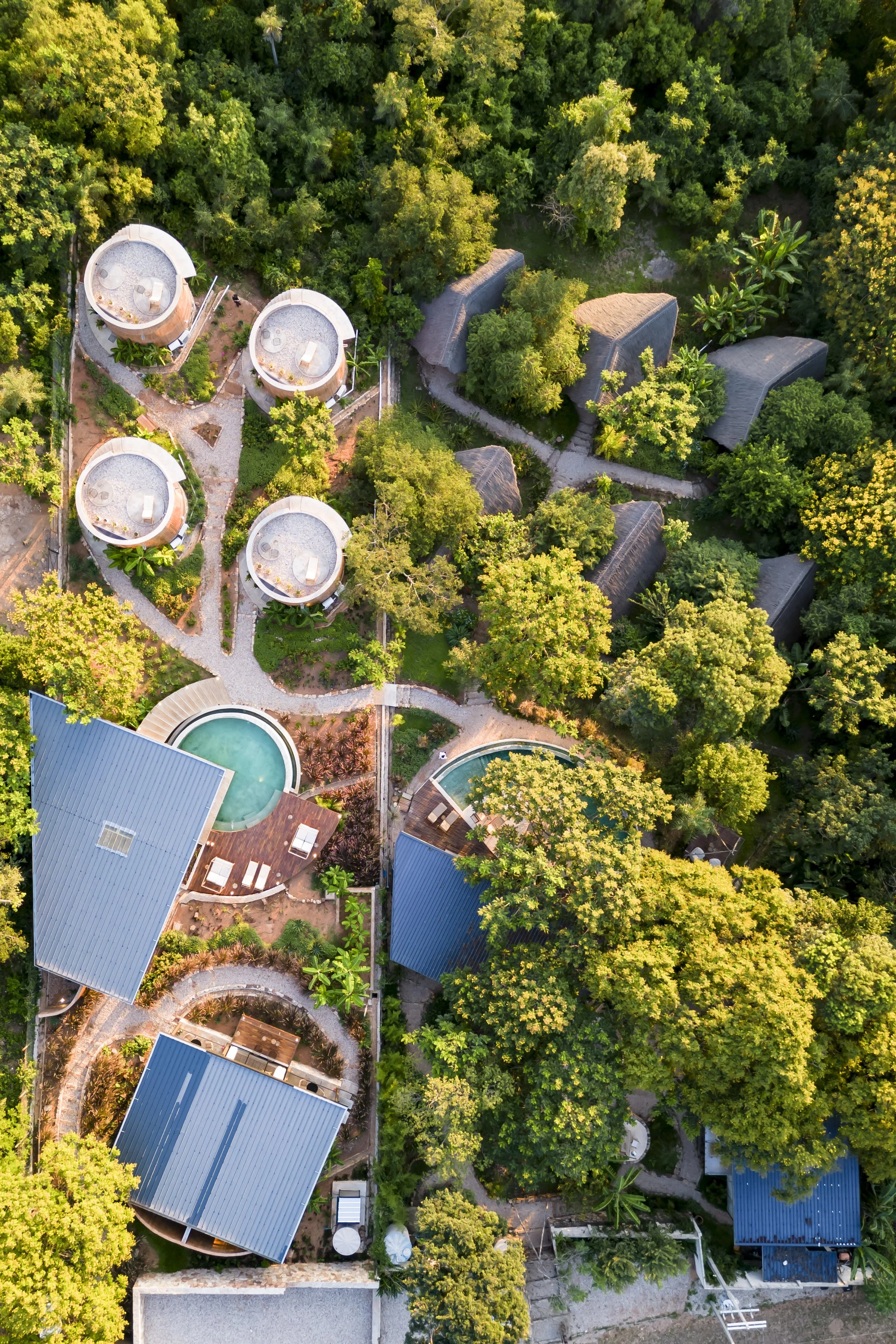 Vista aérea de un complejo turístico rodeado de árboles, con varias cabañas con techos de palma, piscinas y senderos que conectan las construcciones.