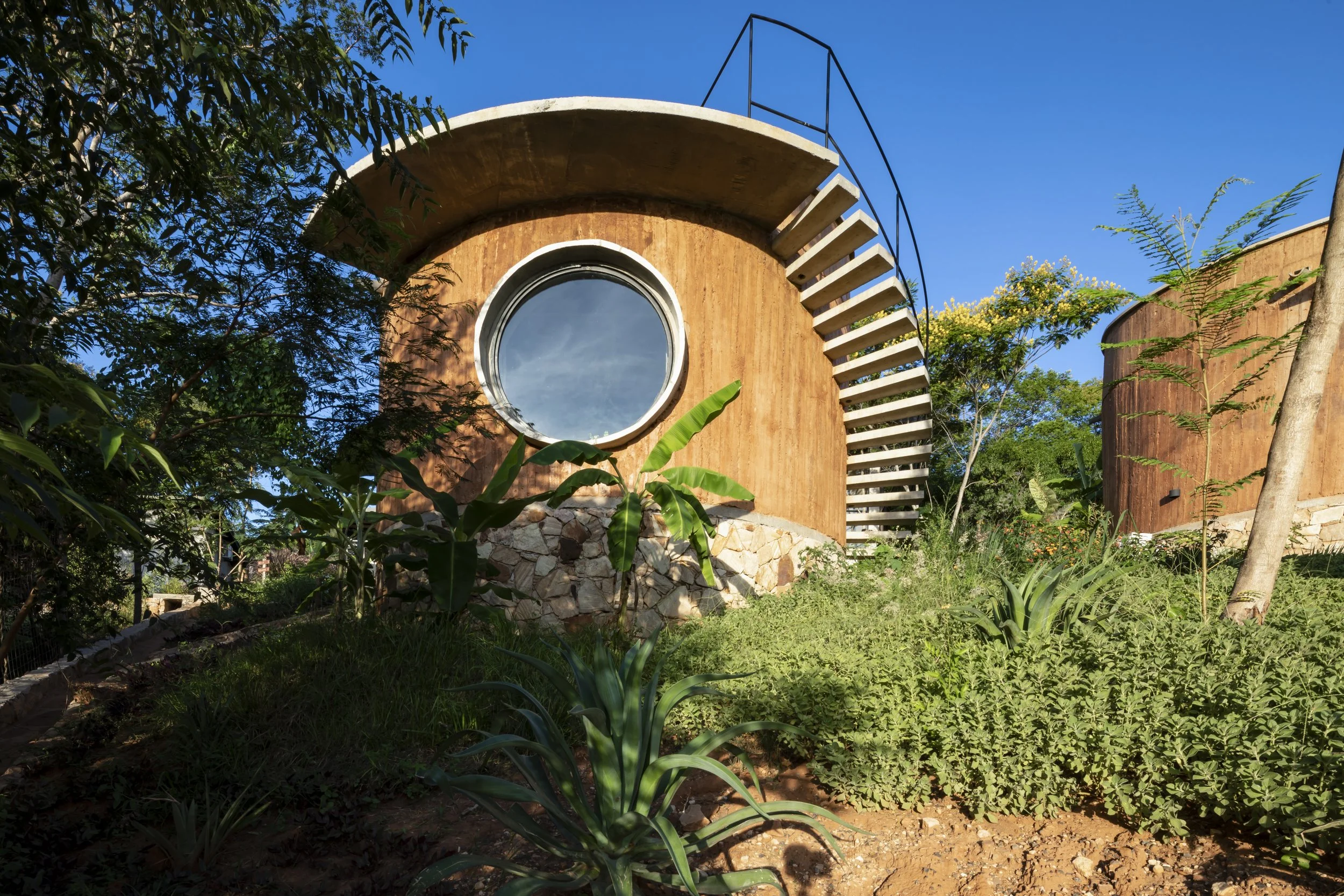 Casa moderna circular de madera y piedra, con una ventana redonda y escaleras en espiral en un entorno natural con plantas y árboles verdes bajo cielo azul.