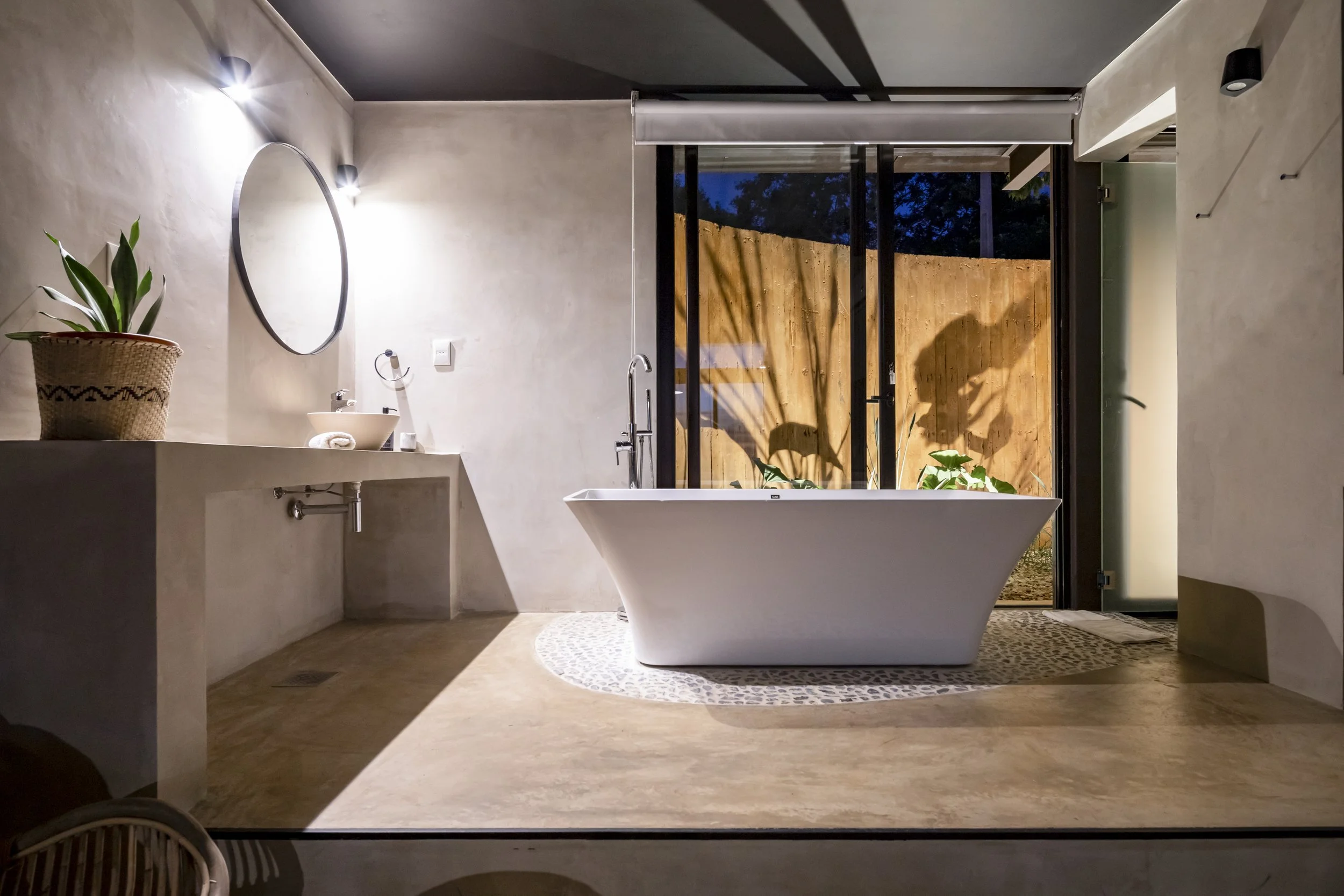 Baño moderno con tina exenta, lavabo y espejo, planta en maceta y vista a un jardín con muro de madera.