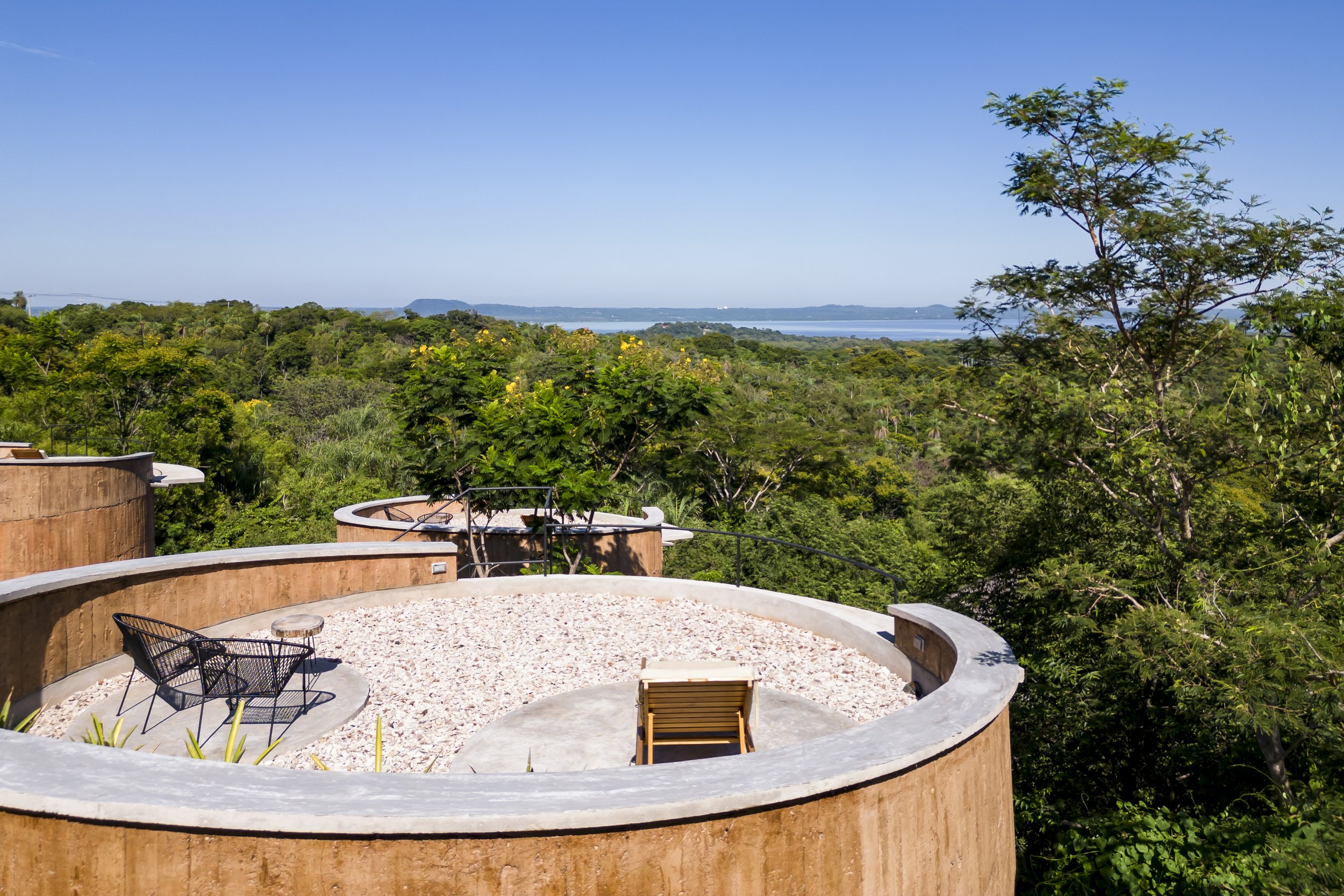 Terraza en una estructura circular con vistas a un paisaje de bosque y agua, con muebles de sillas y una tumbona, rodeada de árboles verdes y un cielo despejado.
