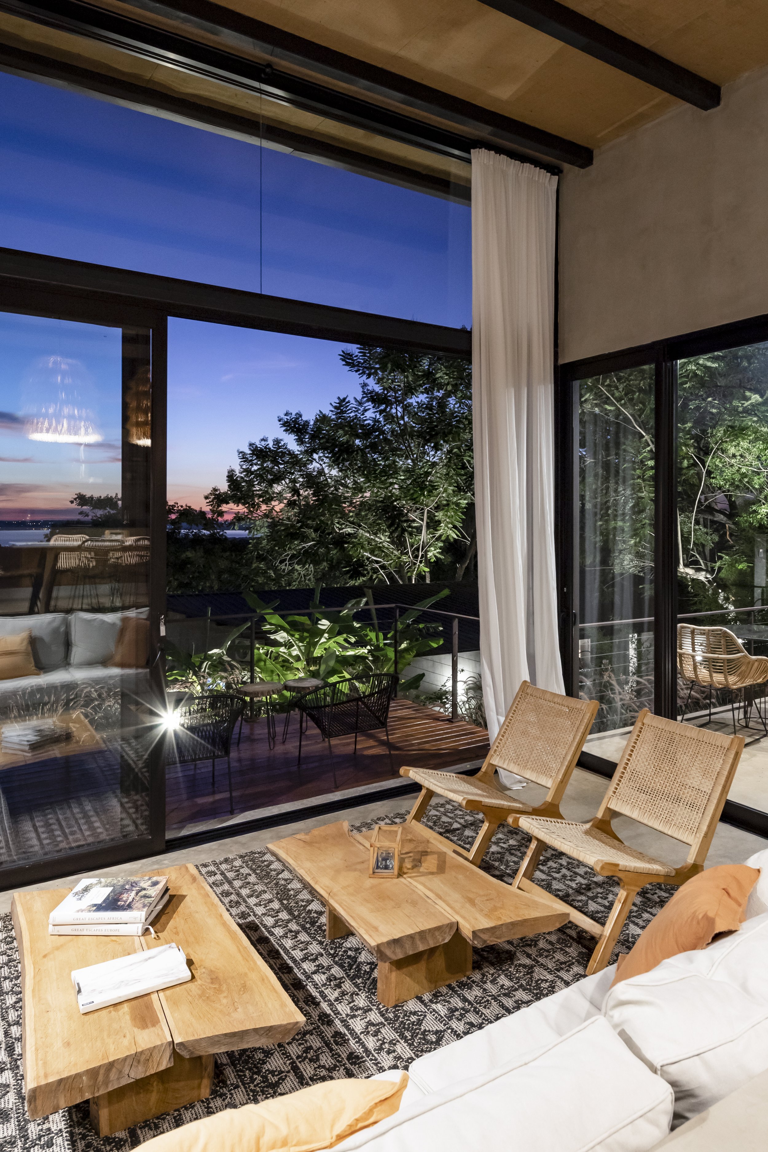 Sala de estar moderna con muebles de madera, sillones de mimbre, una alfombra y vista a un balcón con plantas y árboles, durante el atardecer.