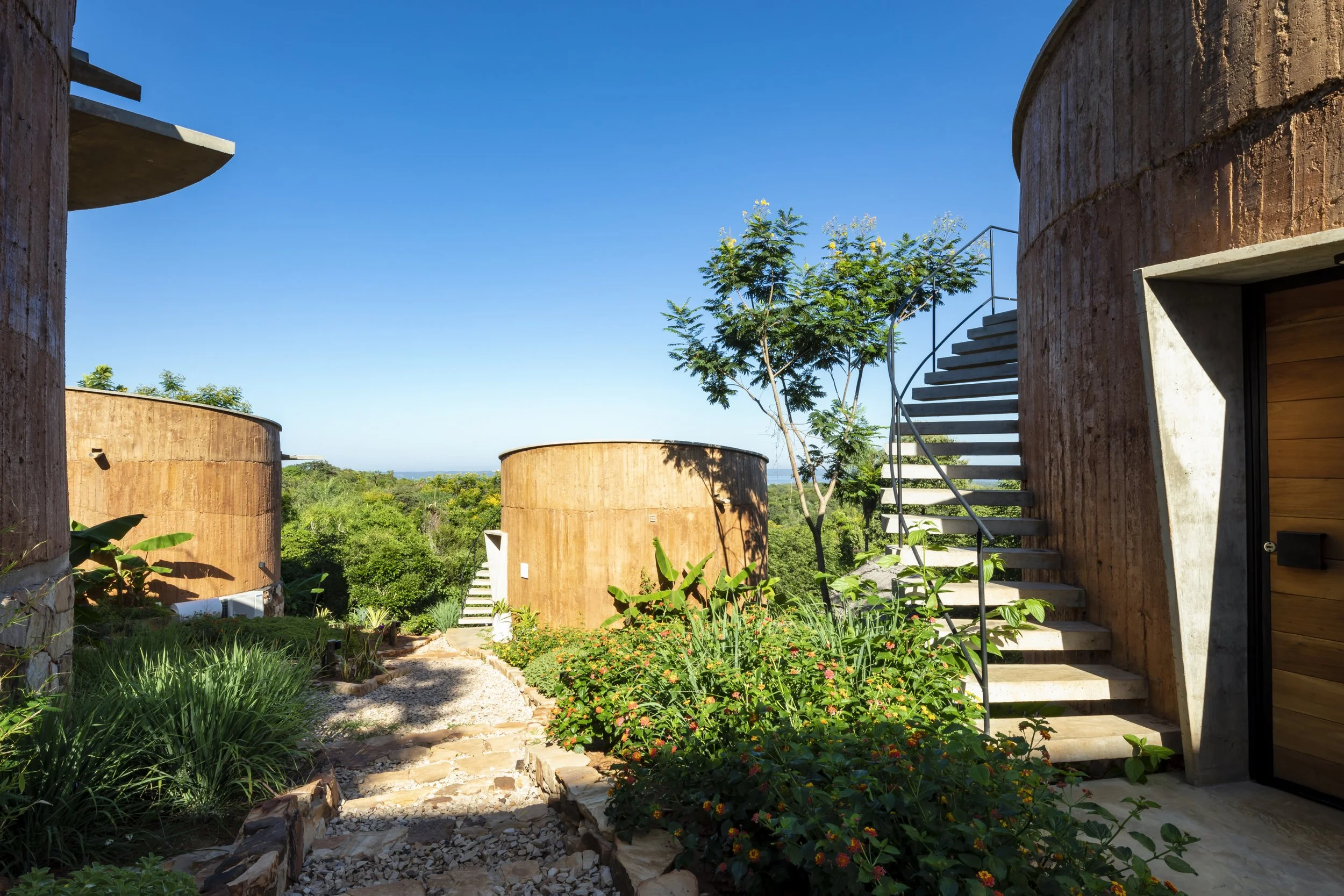 	
PRO ARQUITECTURA PARAGUAY architecture design PROSA
Edificio de arquitectura moderna con paredes de concreto y madera, escaleras exteriores, rodeado de vegetación, árboles y plantas, cielo despejado y paisaje verde en el fondo.