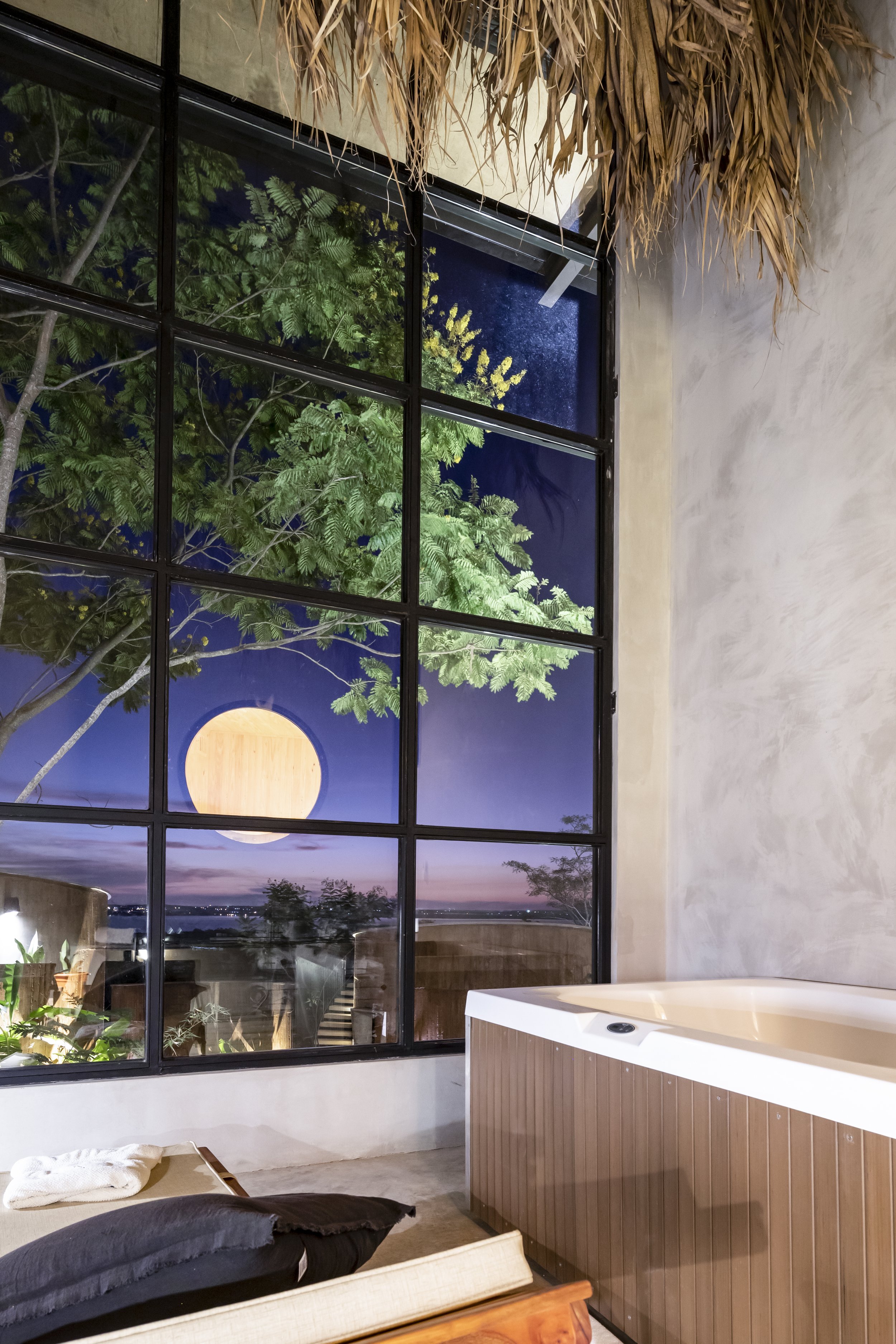 Vista nocturna vista a través de una ventana grande con paisaje con árboles, cielo oscuro y luna llena, vista desde el interior de una habitación con una bañera y un sofá.