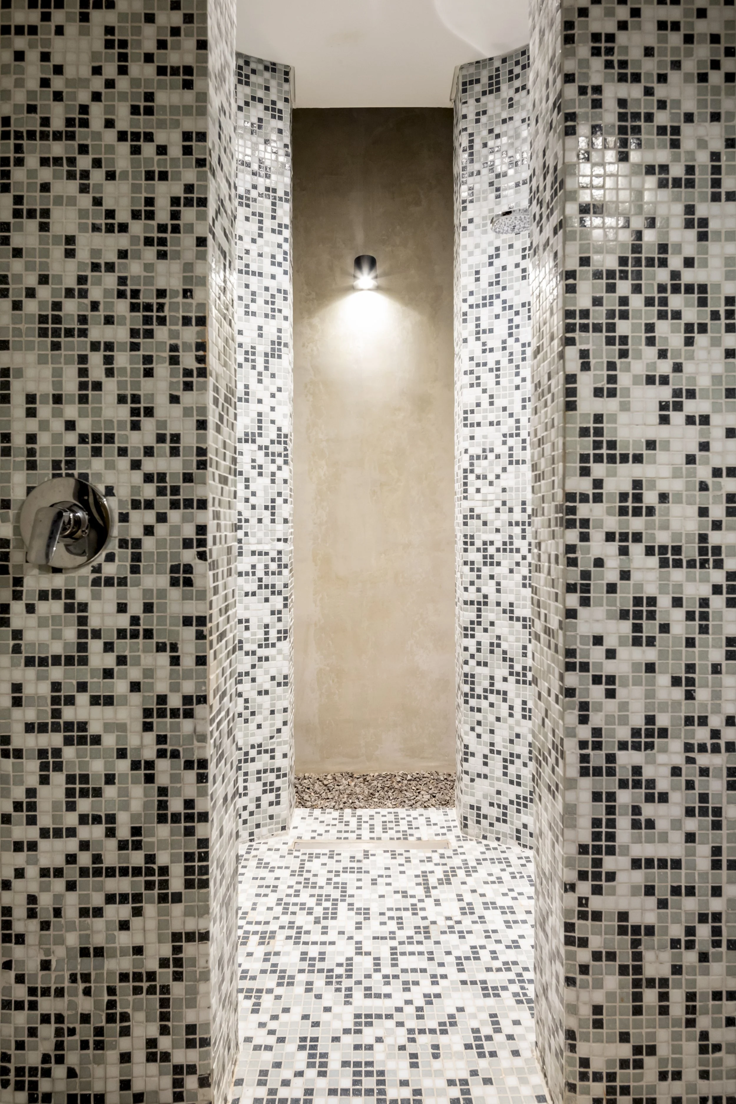 Ducha en baño con paredes y piso de mosaico en tonos blancos, grises y negros, con una iluminación en la pared del fondo.