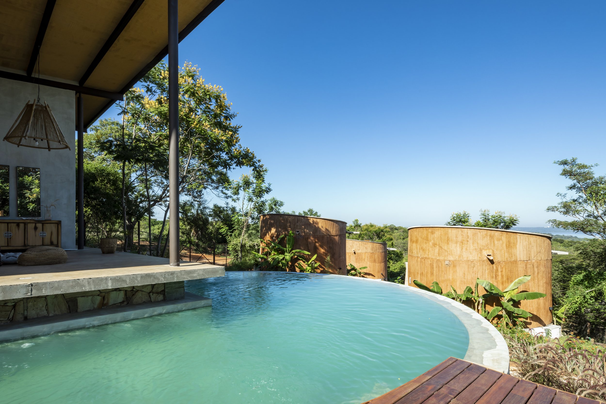 	
PRO ARQUITECTURA PARAGUAY architecture design PROSA
Vista de una piscina de borde infinito en un entorno natural con árboles y construcciones de barro, bajo un cielo azul claro.