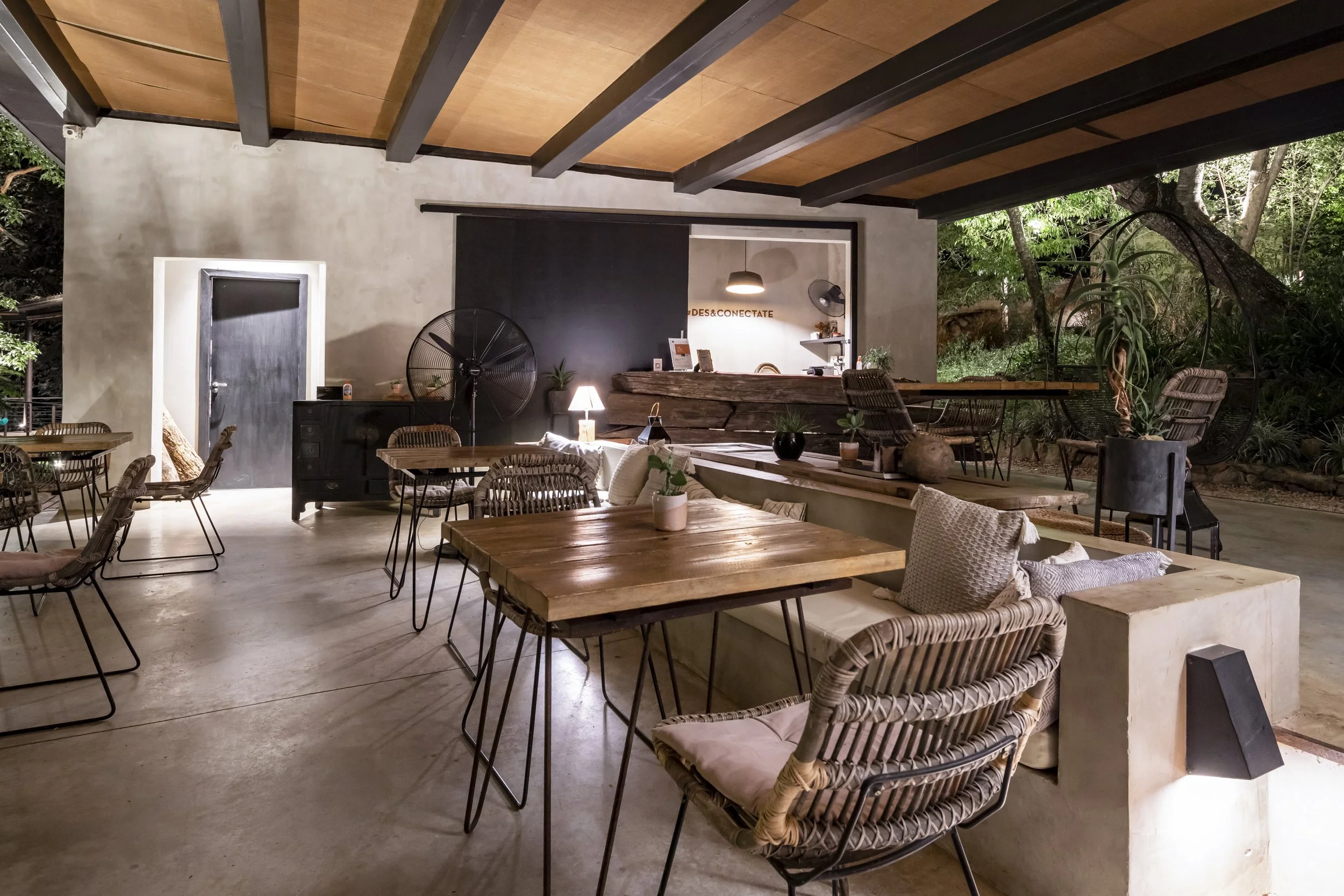 Interior de un restaurante con mesas de madera, sillas de mimbre y decoraciones naturales. Tiene lámparas, plantas y un ambiente acogedor con vista a un jardín exterior.