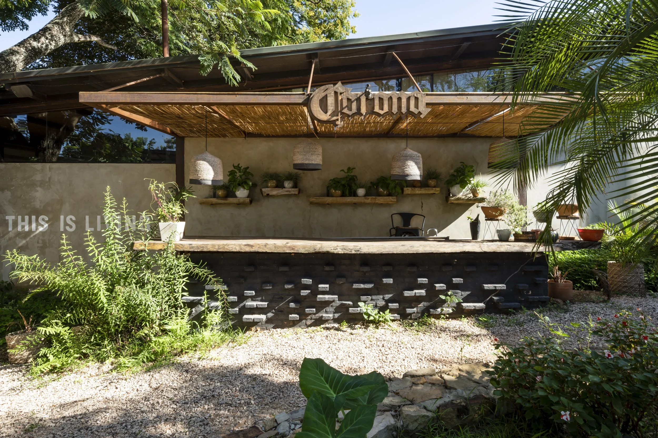 Bar de estilo rústico al aire libre con paredes de concreto y estanterías de madera, decorado con plantas en macetas y lámparas colgantes de fibra, con un cartel que dice 'Corona' y un cartel que dice 'This is LI' parcialmente visible.