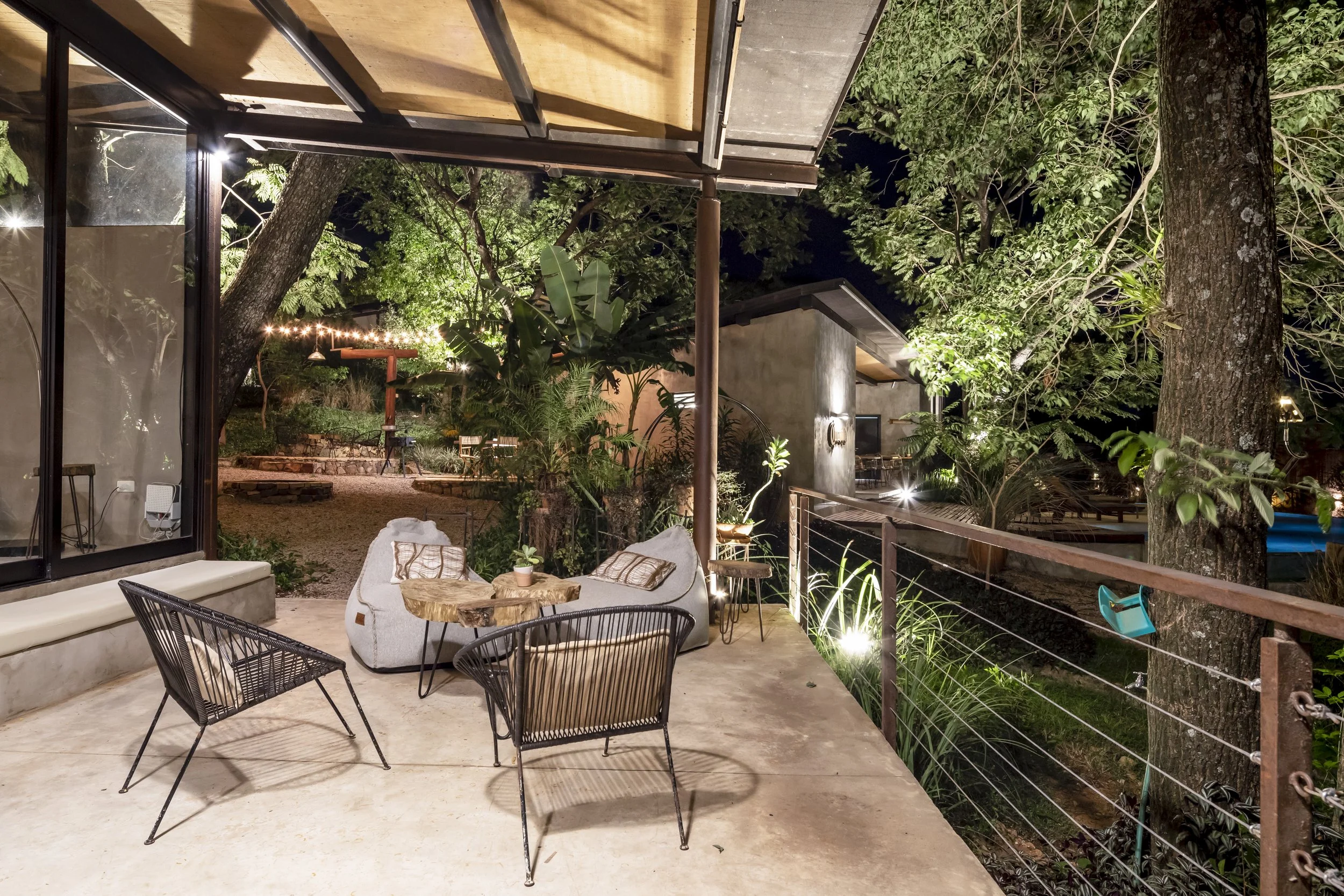 Terraza exterior con sillones, mesa de madera, árboles grandes, iluminación nocturna y jardín en el fondo.