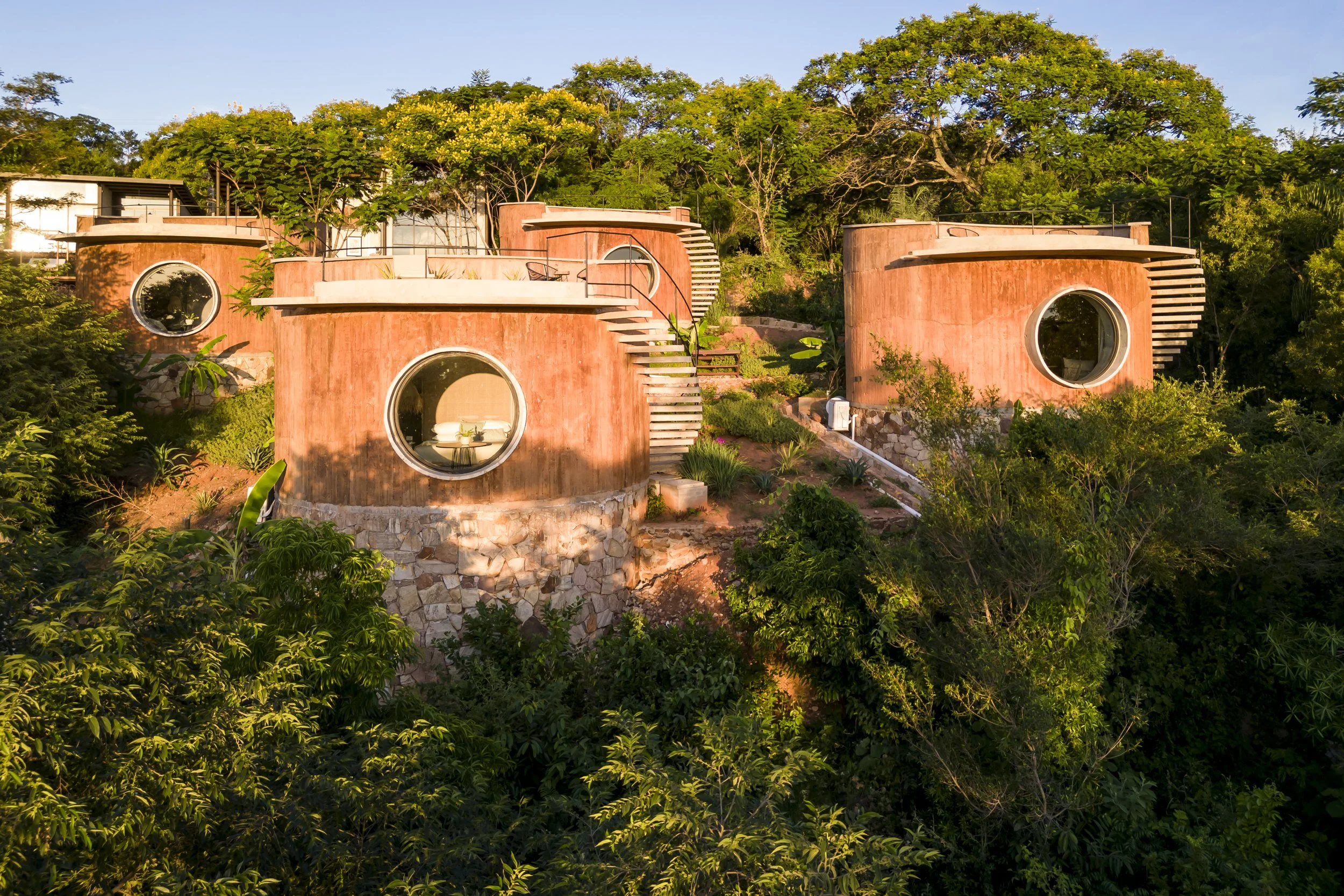 Casas de diseño innovador con paredes de color terracota y ventanas redondas, situadas en una colina rodeada de vegetación y árboles verdes.