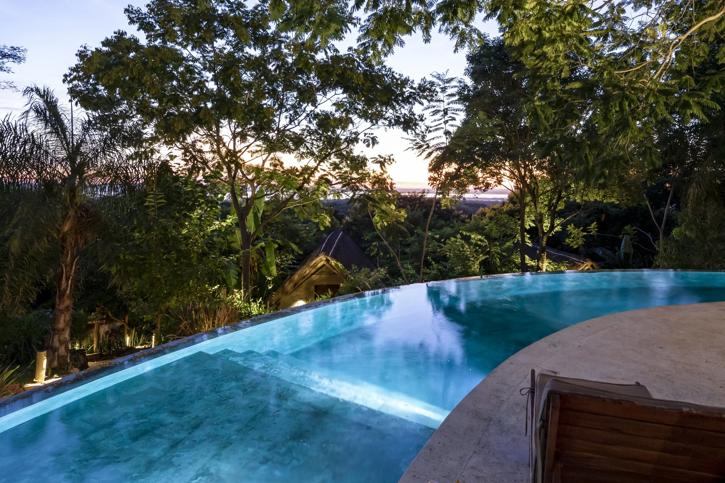 Piscina con vista a la naturaleza, árboles y un atardecer en el fondo.