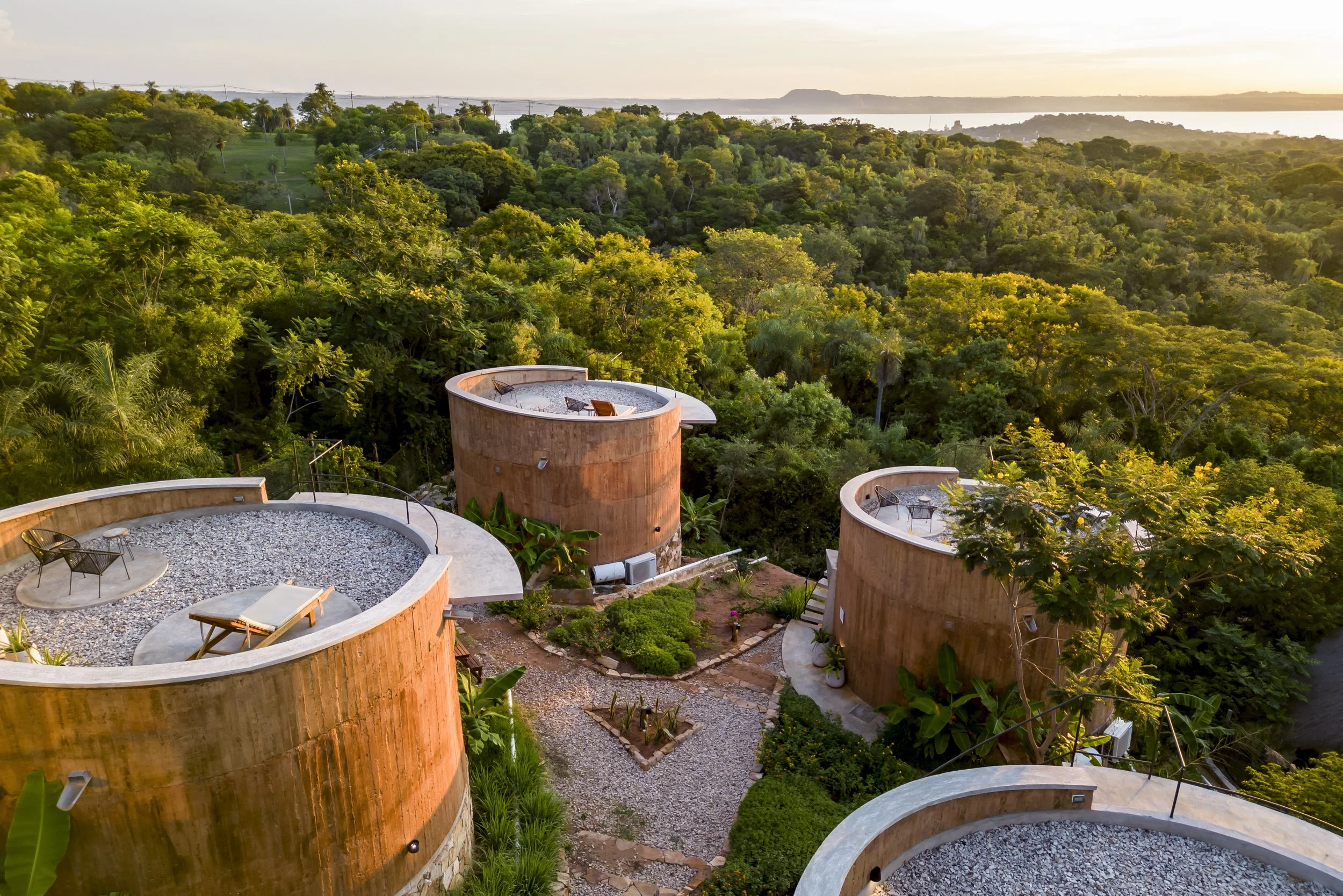 Edificio de diseño circular con terrazas en medio de un área boscosa, vista hacia un lago o río con colinas al fondo, atardecer.