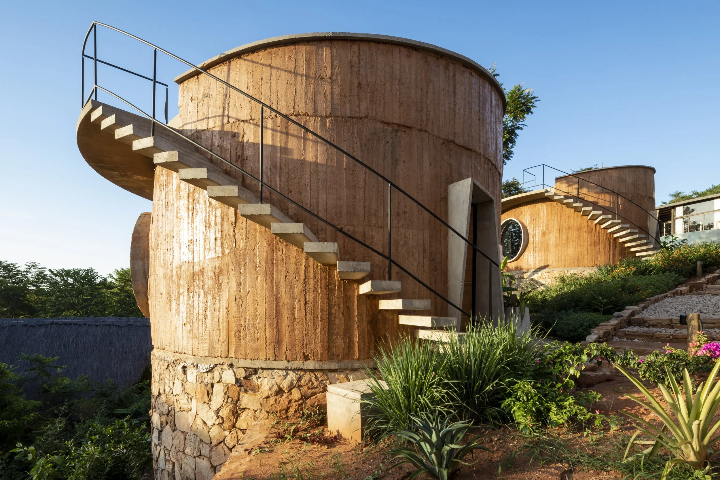 Edificio moderno con paredes de ladrillo y escaleras de caracol exteriores, rodeado de vegetación y plantas en un paisaje con cielo despejado.