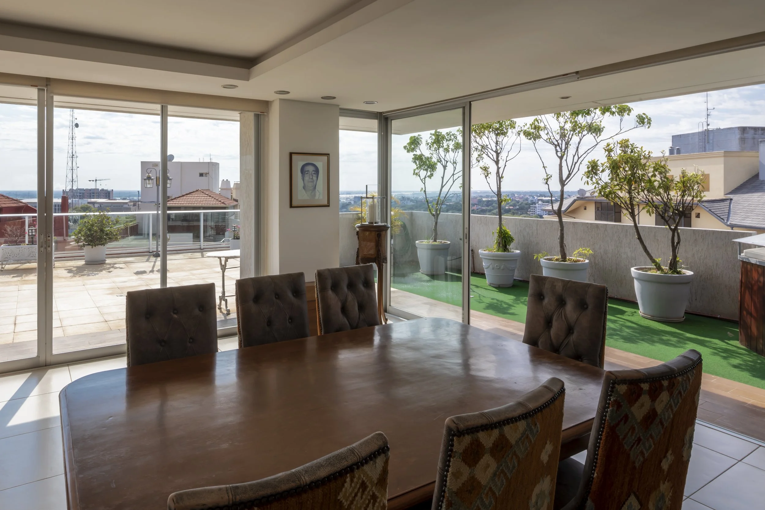 Sala de juntas con mesa de madera y sillas tapizadas, vista a un balcón con macetas y plantas verdes, ventanas grandes con vista a la ciudad, decoración sencilla con retrato en la pared y una lámpara.