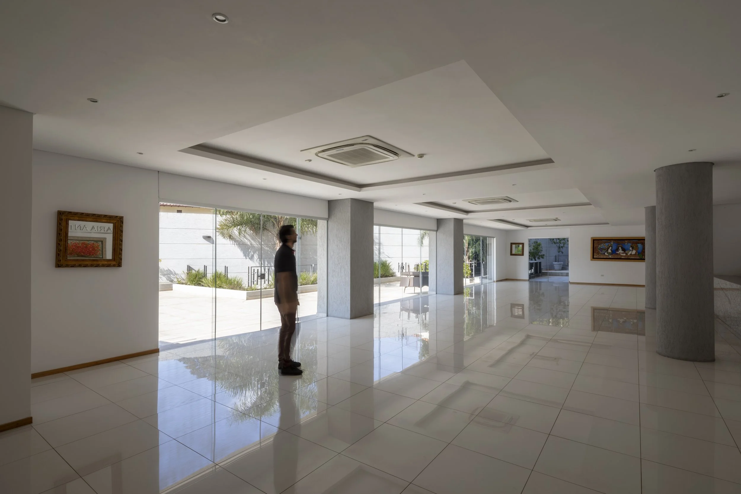 Interior de un vestíbulo amplio con paredes blancas, varias ventanas grandes, columnas y cuadros en las paredes. Una persona observa hacia afuera a través de las ventanas y el suelo es de baldosas brillantes.