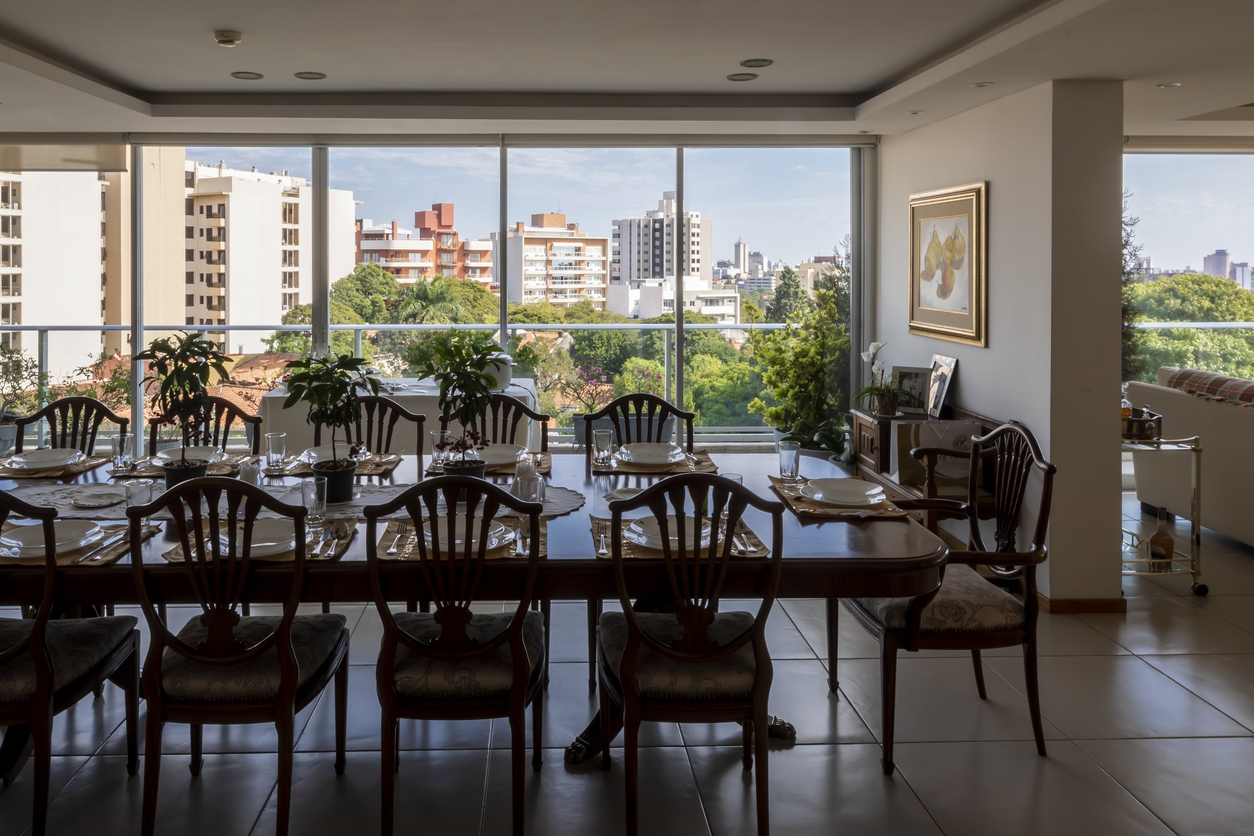 Comedor con mesa lista para cenar, grandes ventanas con vista a edificios y árboles, decoraciones con plantas y pinturas.