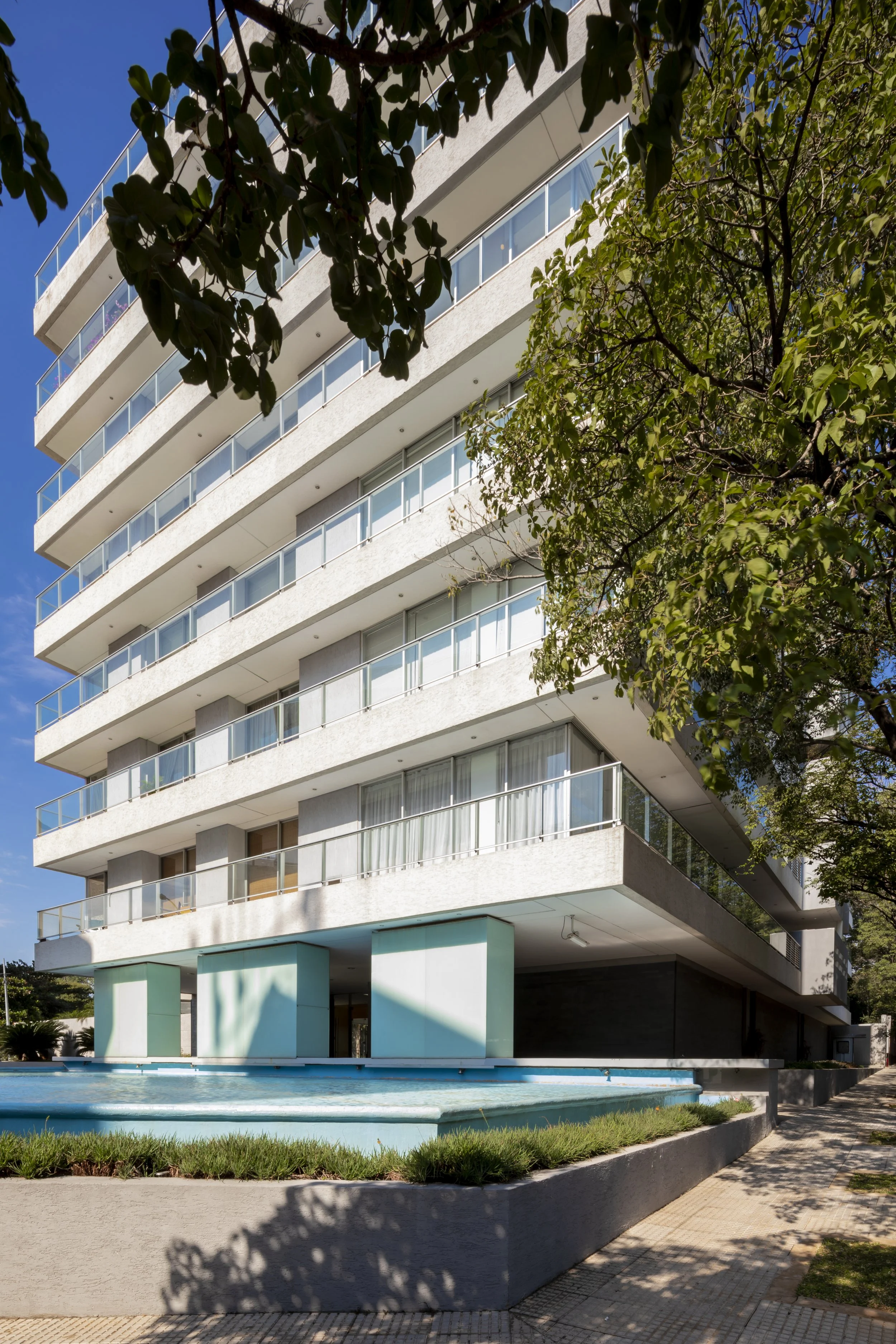 PRO ARQUITECTURA PARAGUAY arquitectos design architecture PROSA
Edificio moderno de varios pisos con balcones de cristal, rodeado de árboles y una fuente en el primer plano, en un día soleado.