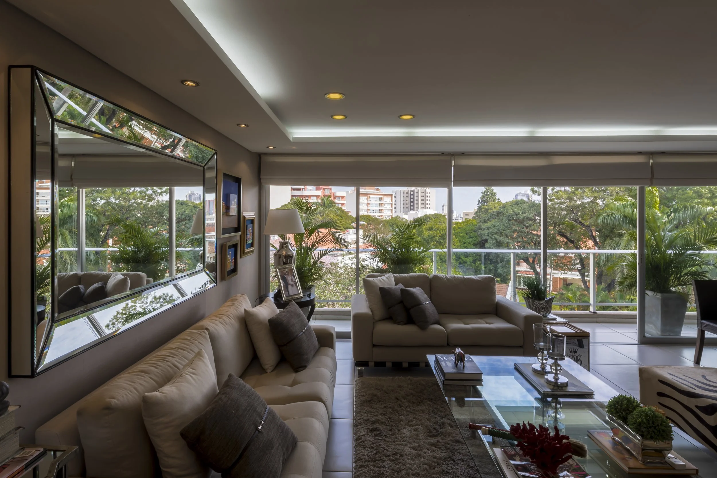 Sala moderna con sillones beige, una mesa de centro de cristal, plantas en la terraza y vistas a la ciudad y árboles.