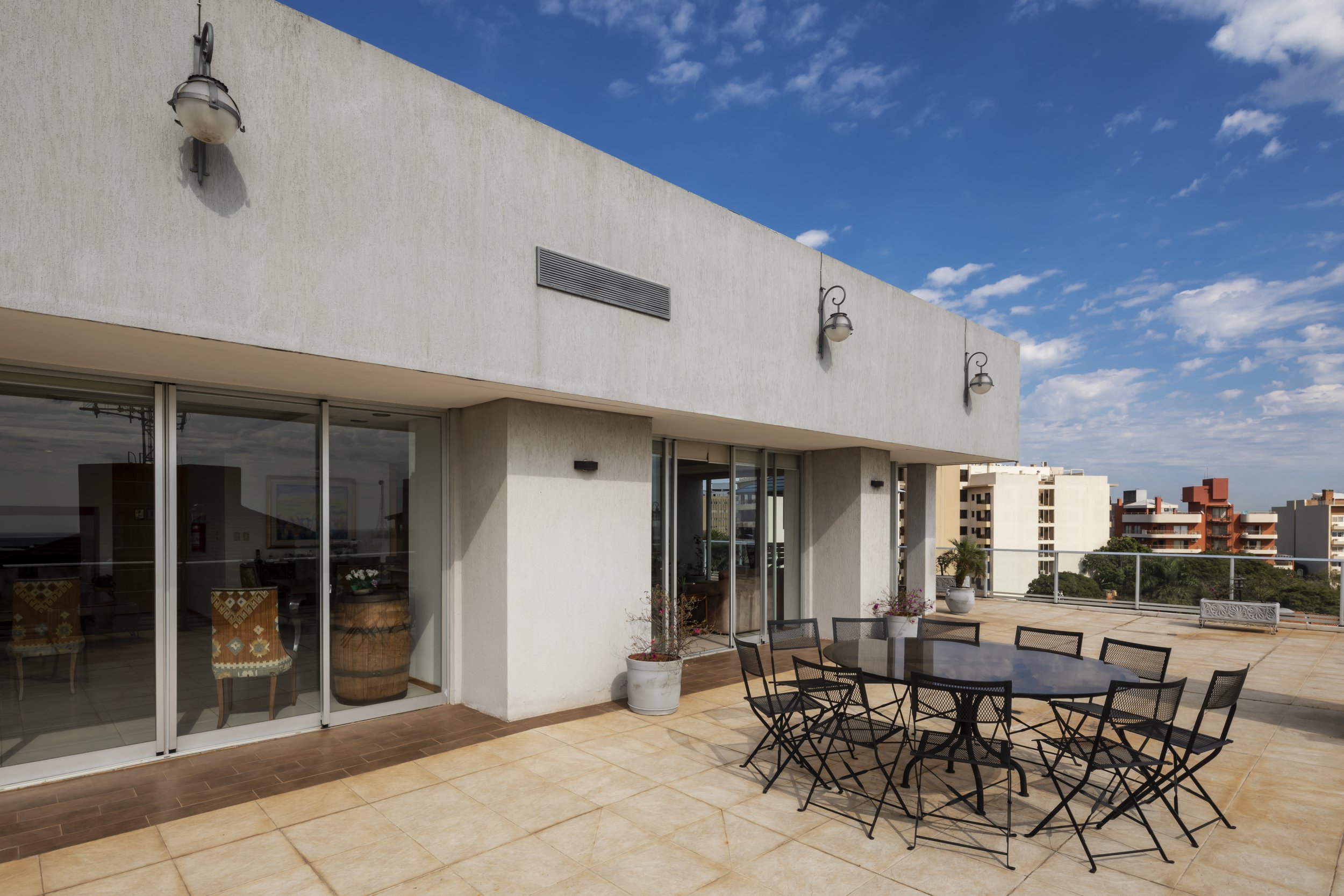 Terraza de edificio con mesa y sillas de metal, plantas en macetas blancas, paredes blancas y ventanas de vidrio, ciudad de fondo con edificios, cielo despejado con nubes.
