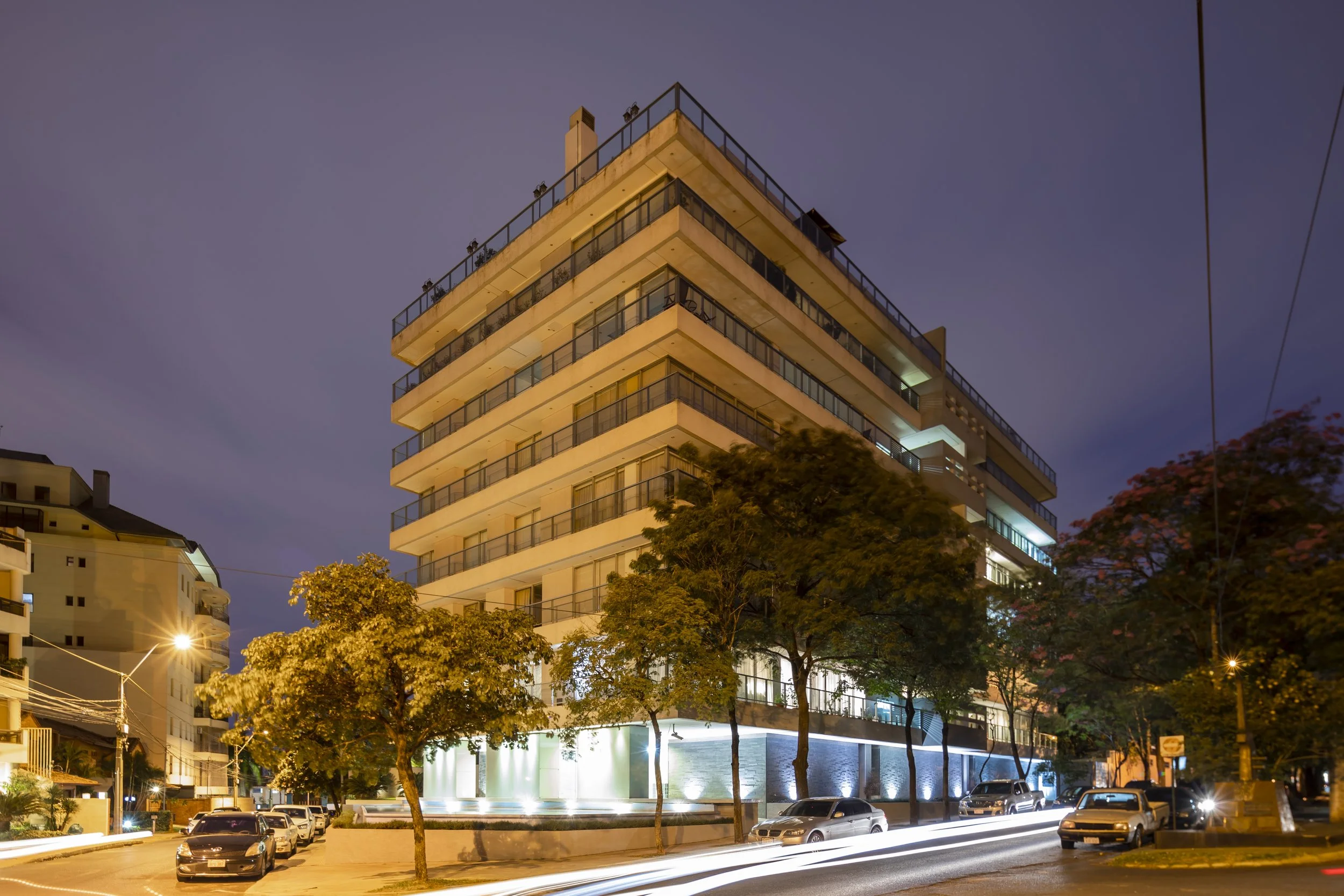 Edificio moderno de varios pisos en una ciudad durante la noche, con árboles y varios autos estacionados frente a la calle iluminada.
