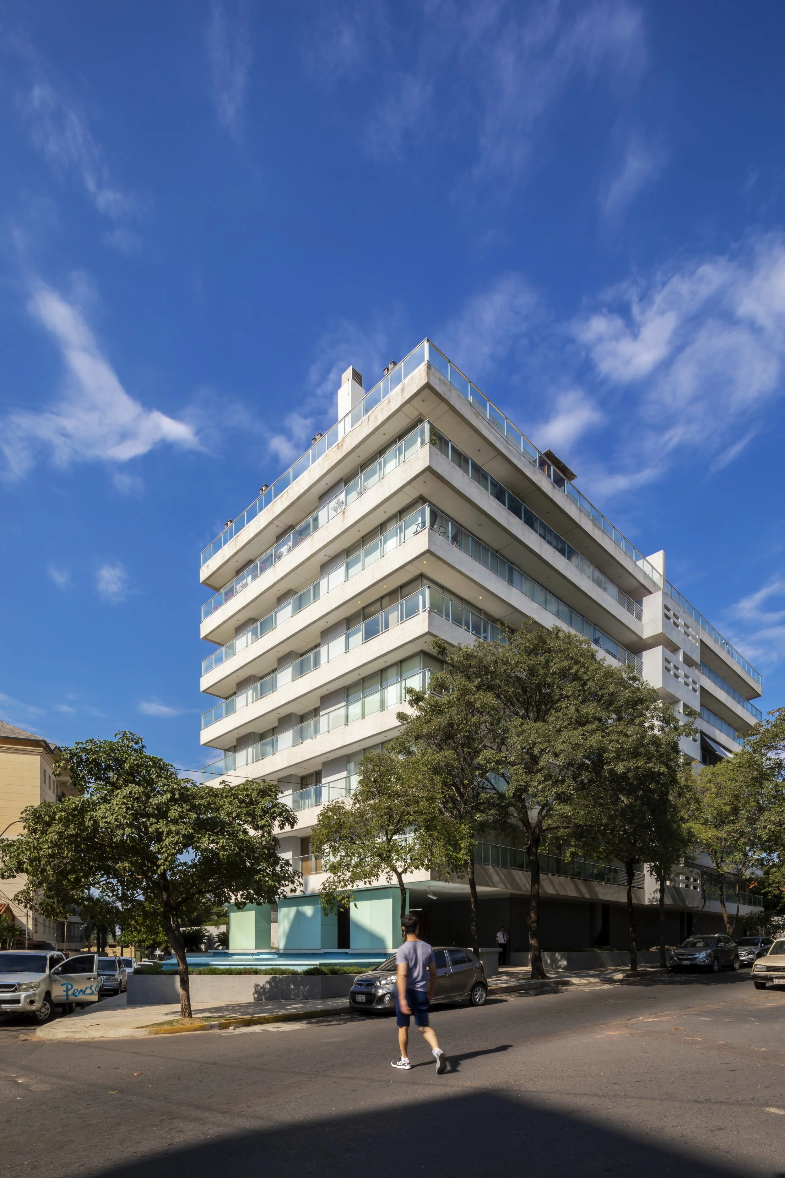 PRO ARQUITECTURA PARAGUAY arquitectos design architecture PROSA
Edificio moderno de varias plantas con terrazas y balcones, rodeado de árboles y coches estacionados, bajo un cielo azul con nubes finas.