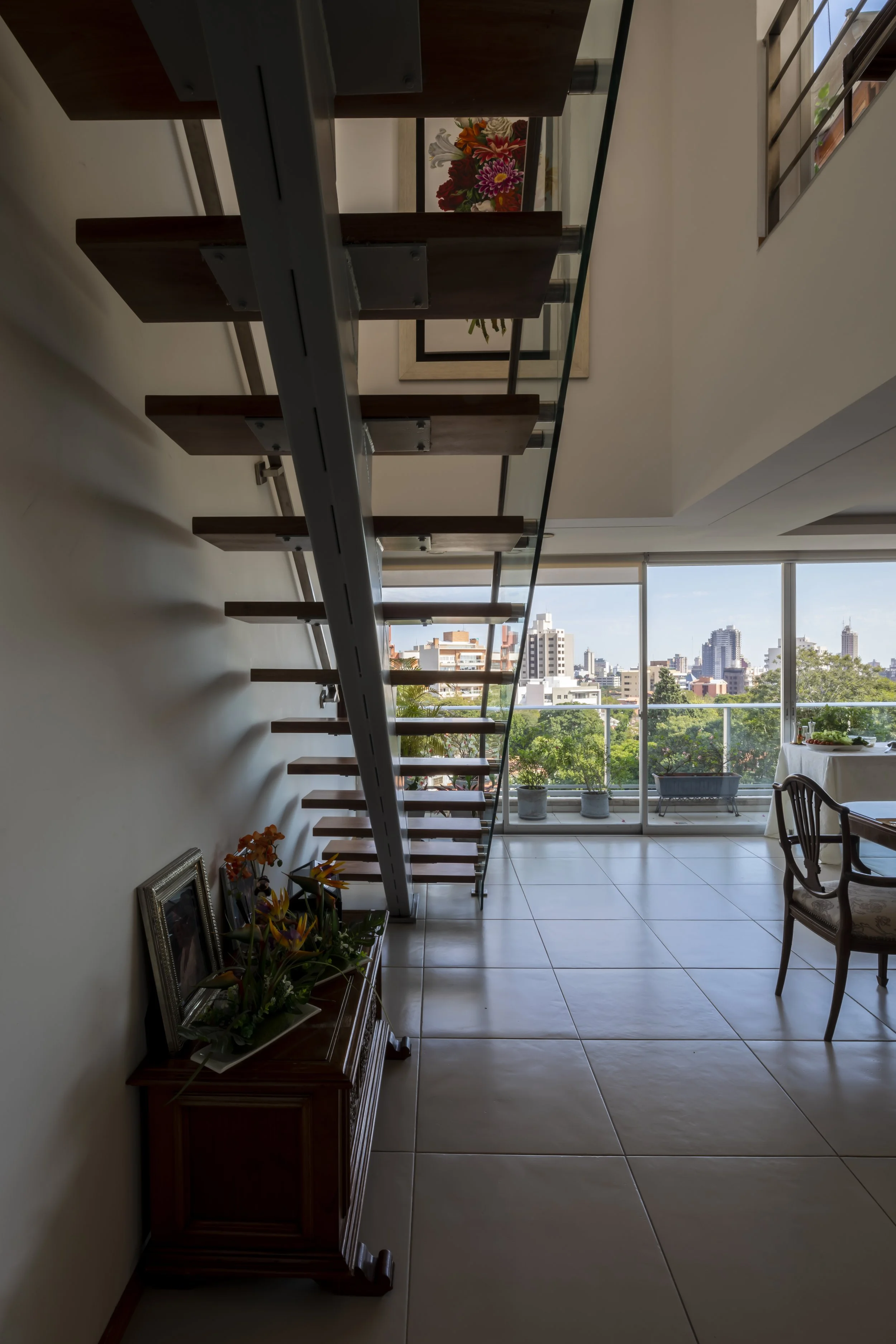 Interior de una sala con una escalera moderna de madera y metal, un piso de baldosas grandes y un ventanal que ofrece vista a un paisaje urbano con edificios altos y áreas verdes, decorada con cuadros y plantas.