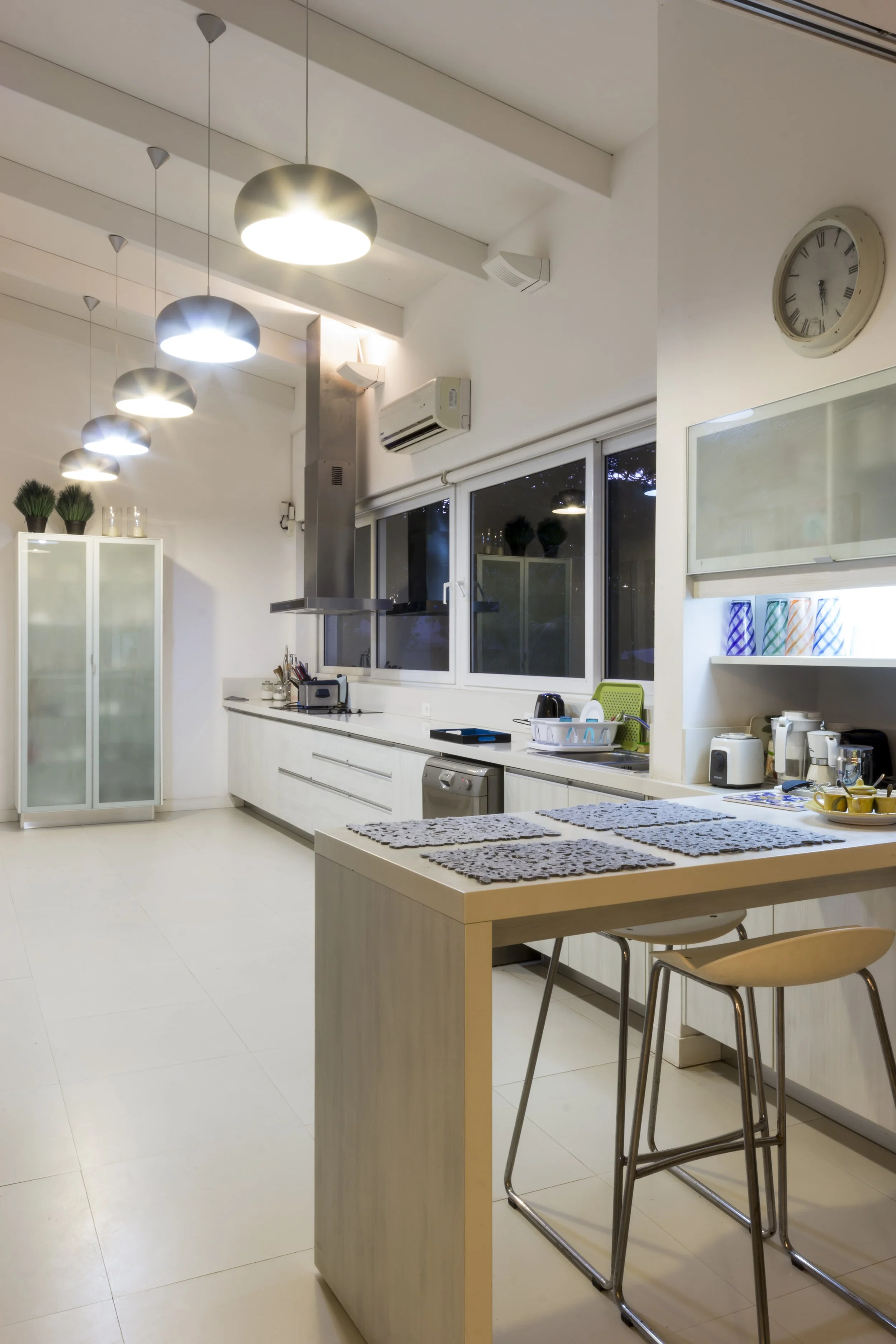 Cocina moderna y luminosa con lámparas colgantes, electrodomésticos blancos, ventanas grandes, y una mesa con manteles en color gris.