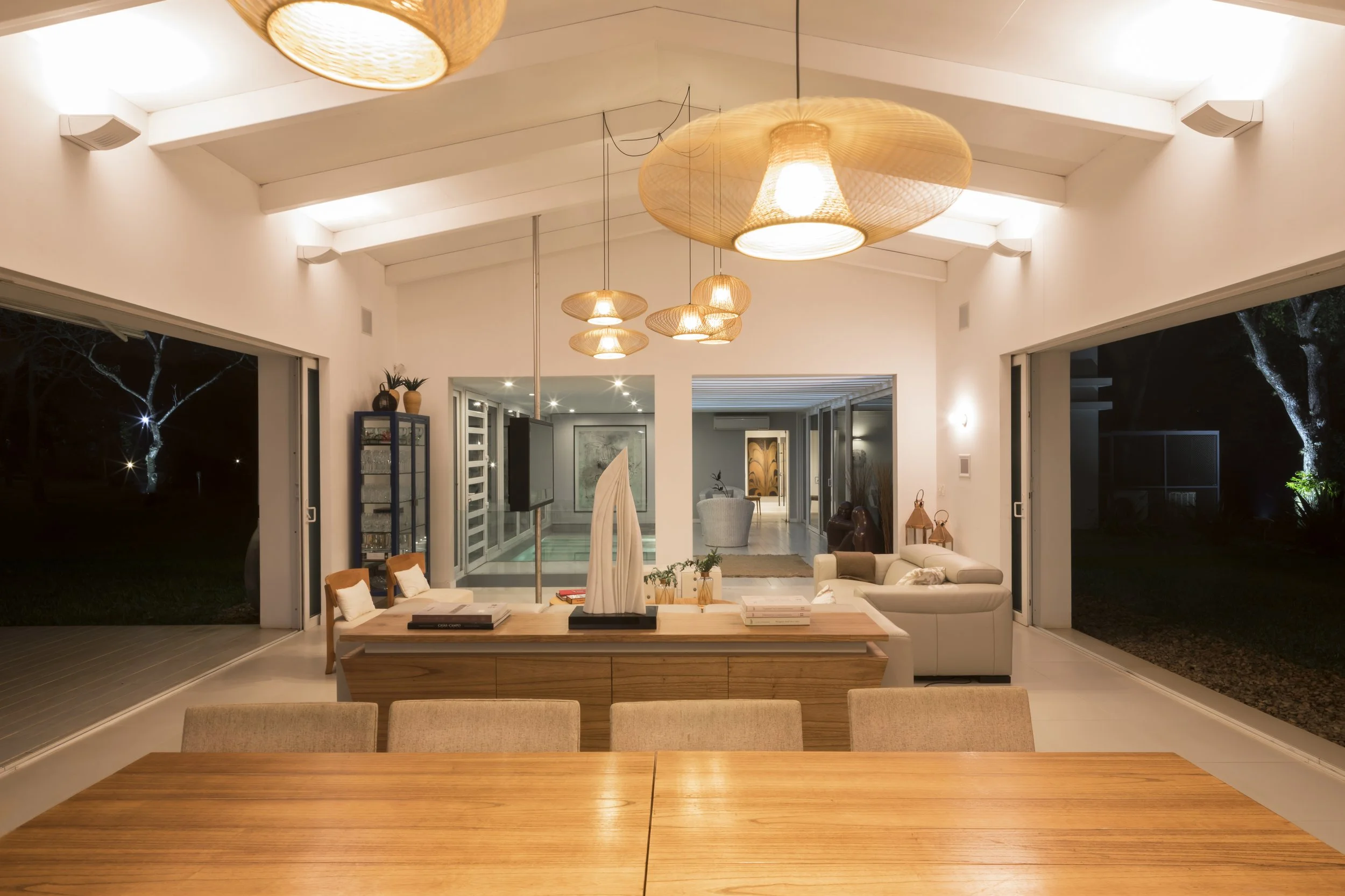 Sala de estar moderna con muebles blancos y de madera, iluminada por lámparas de techo de mimbre, con puertas corredizas abiertas hacia el exterior en la noche.