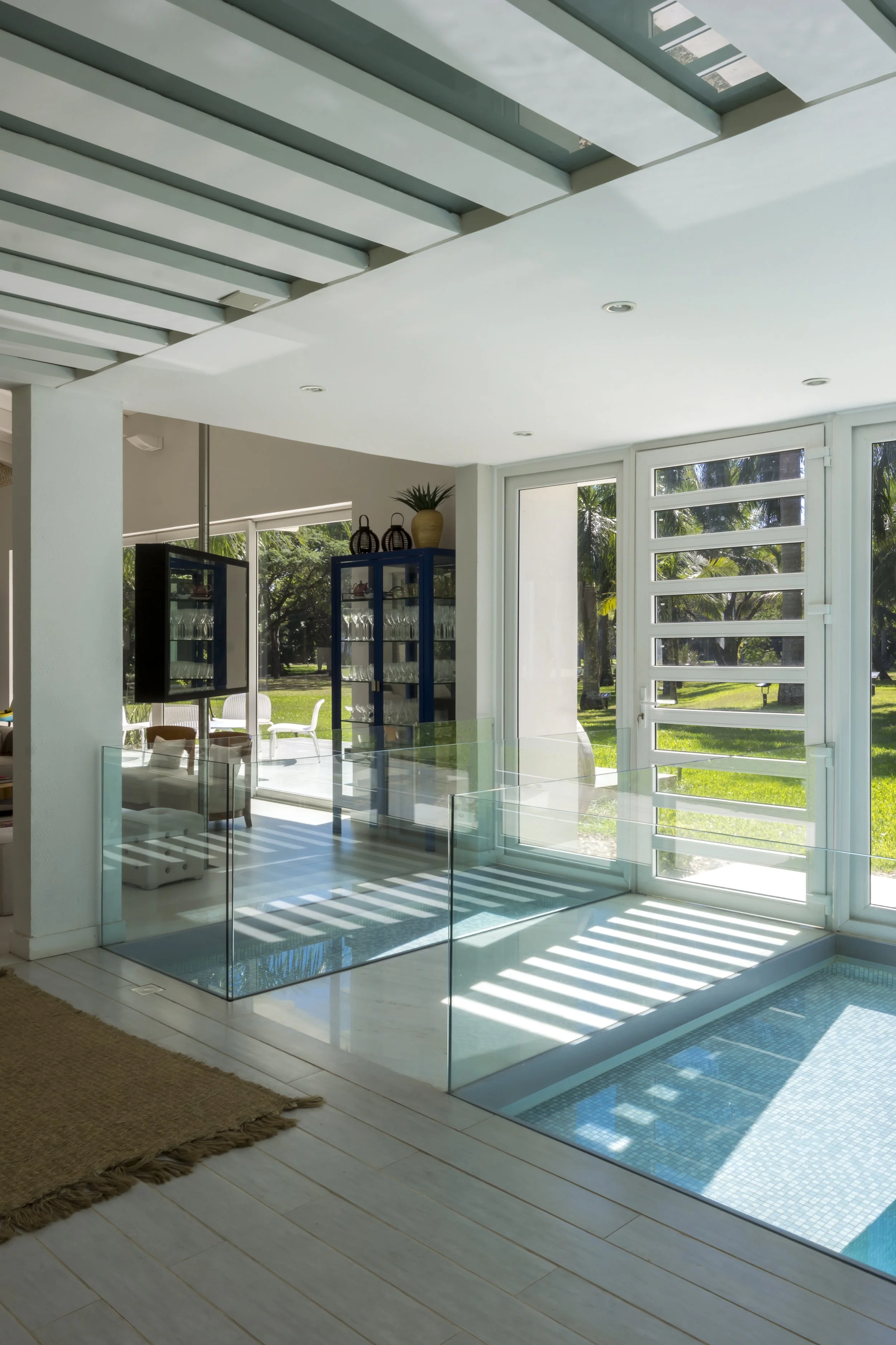 Interior de una casa con ventanas grandes, un área con piscina pequeña rodeada de acrílico, muebles y decoración moderna, y vista hacia un jardín con árboles.
