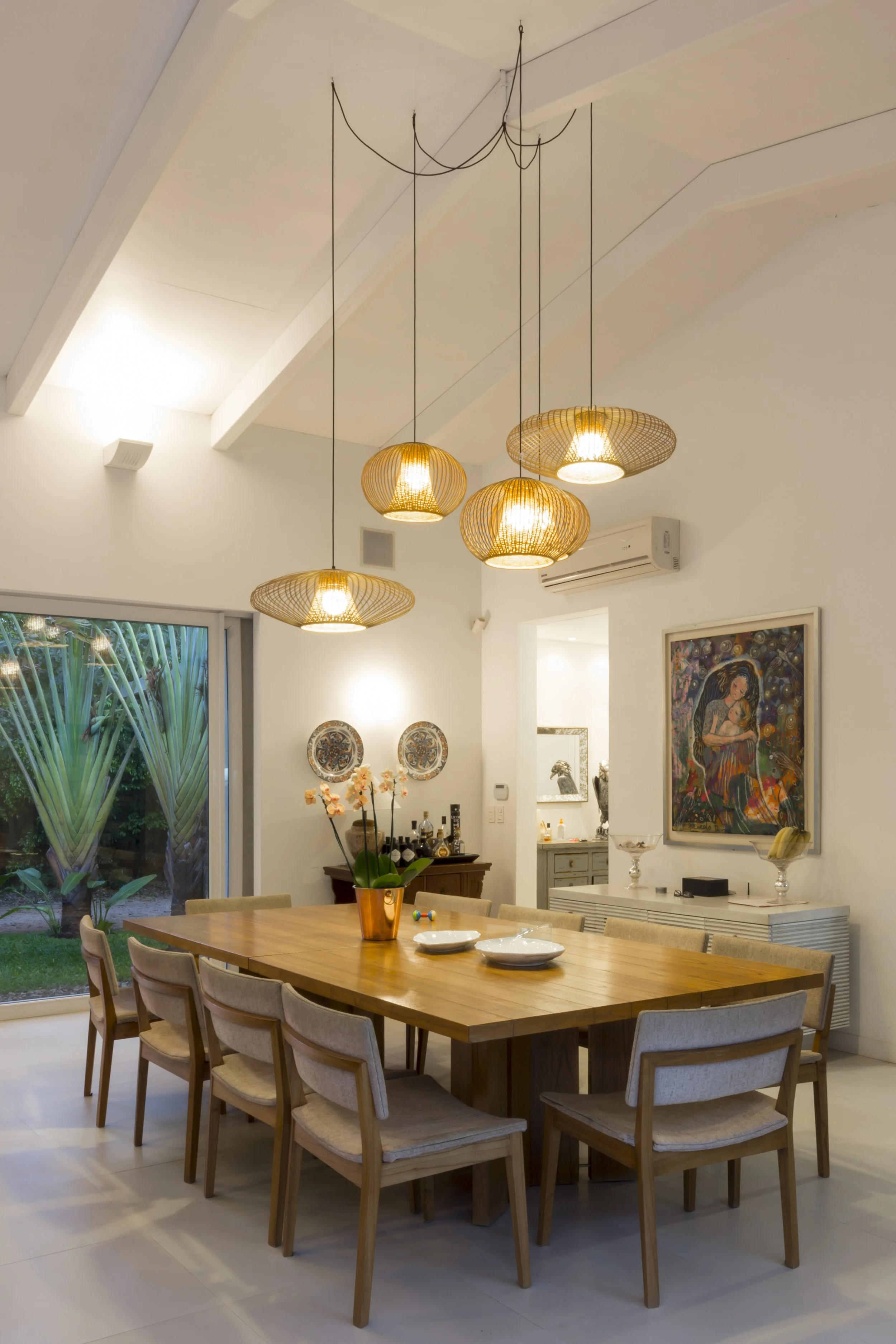 Sala de comedor con mesa de madera rodeada de ocho sillas, lámparas colgantes de mimbre sobre la mesa, ventana grande con vista a un jardín con plantas verdes, decoraciones en las paredes y un mueble con objetos decorativos.