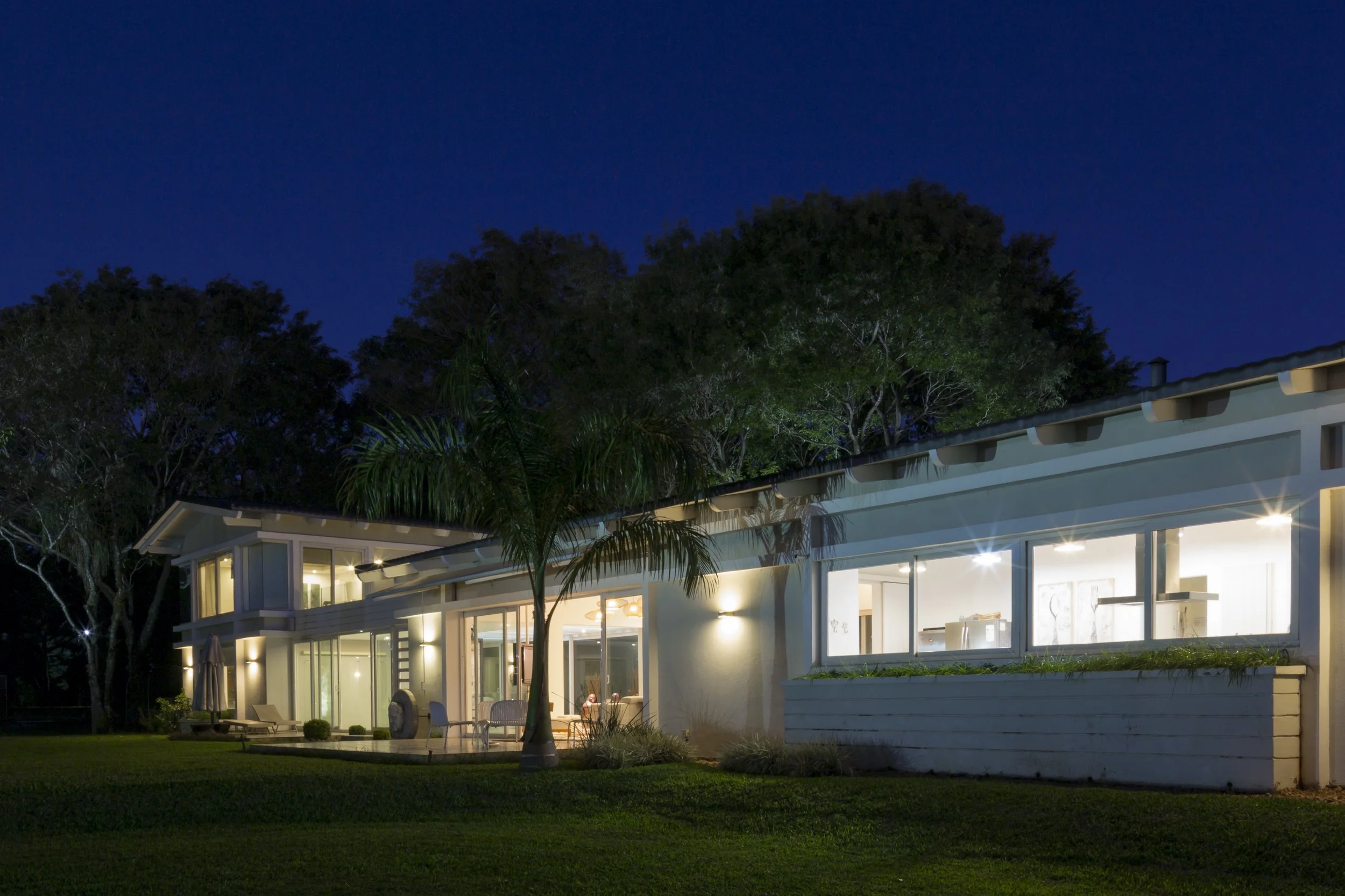 Casa moderna iluminada por la noche con techo inclinado y grandes ventanas, rodeada de árboles y césped.