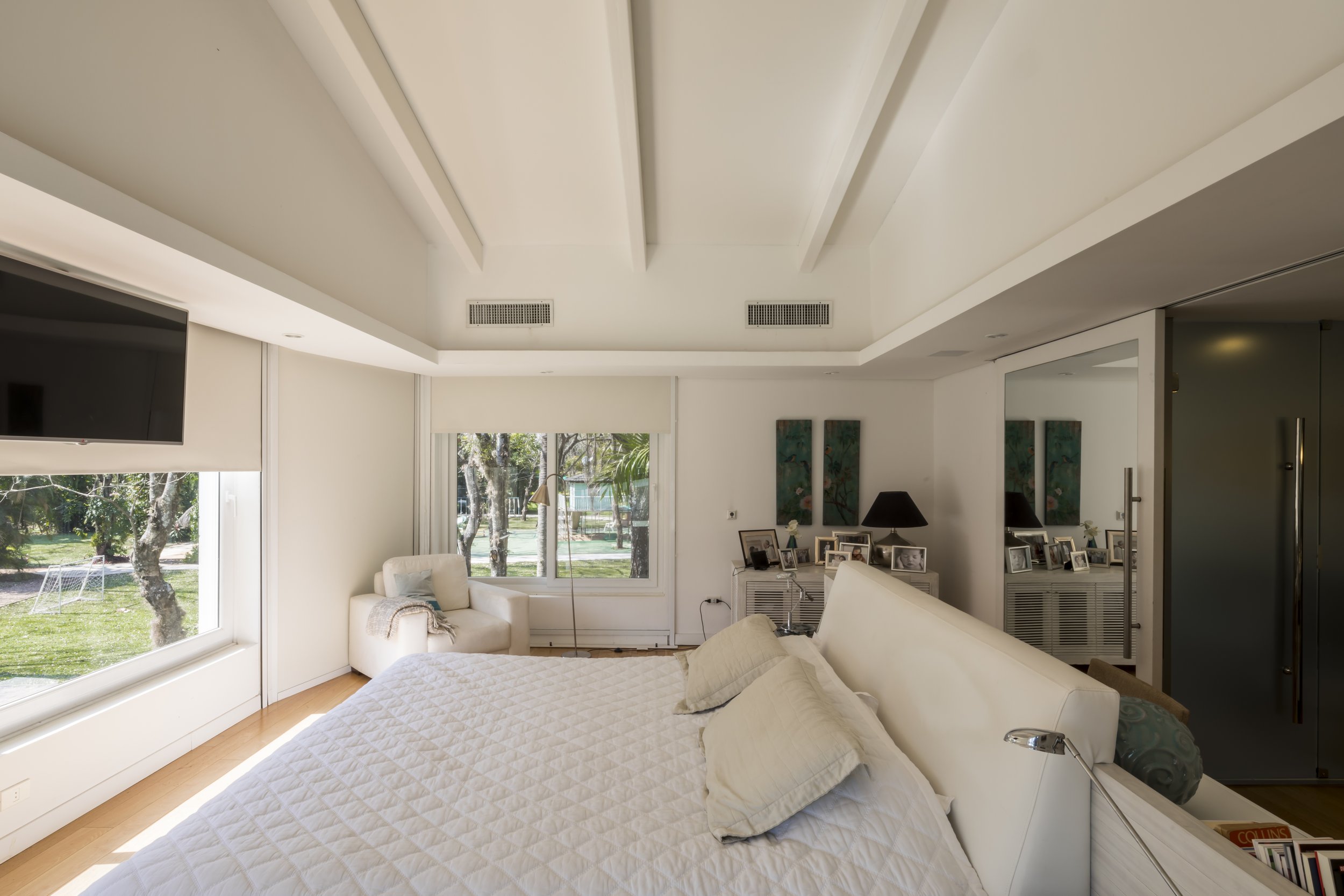 Habitación moderna con cama grande, paredes blancas, ventanas grandes con vista a un jardín, y decoraciones con fotografías y cuadros en la pared.