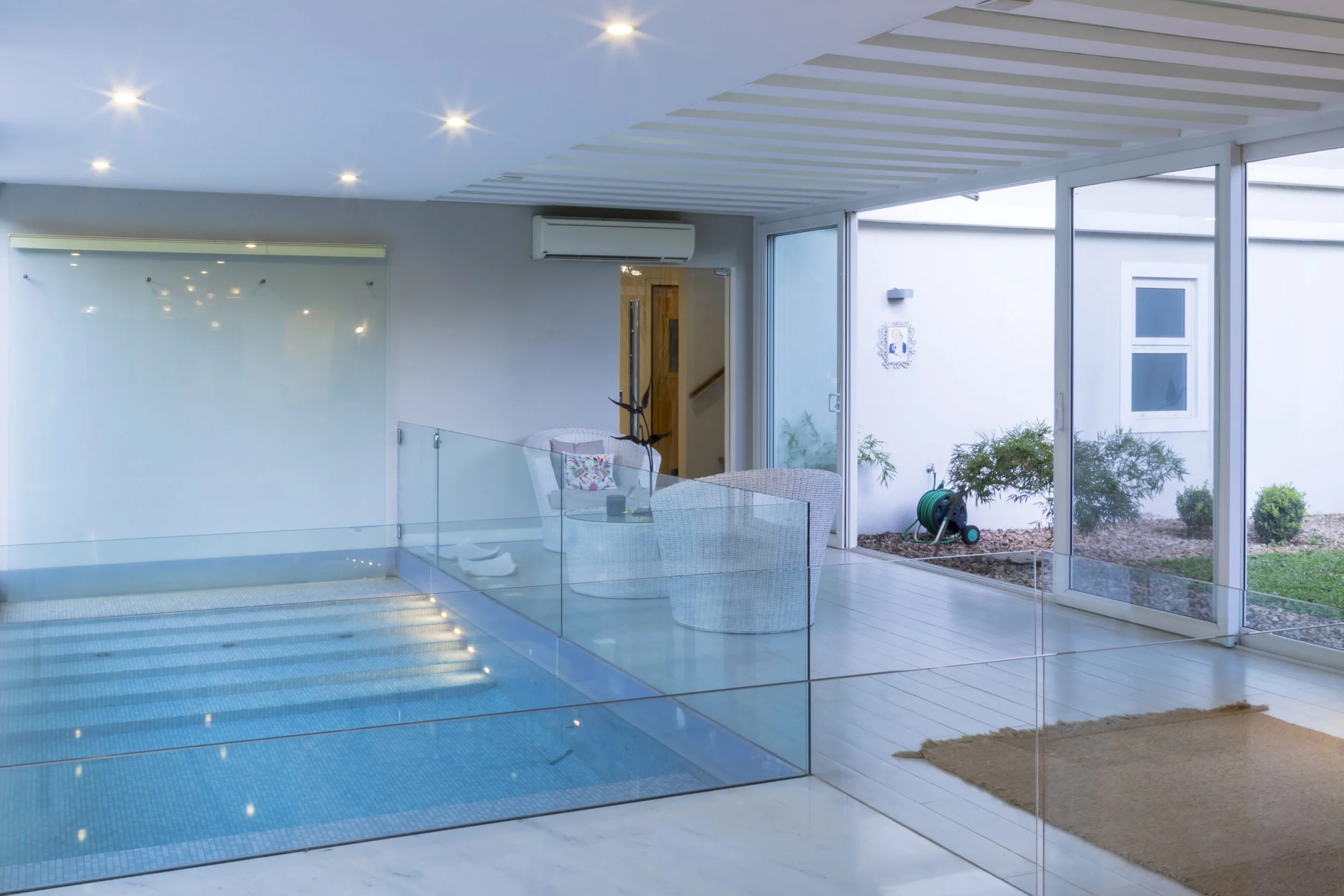 Interior de una propiedad moderna con piscina cubierta, muebles blancos, plantas en el exterior, segadora de césped y ventanas grandes que permiten la entrada de luz natural.