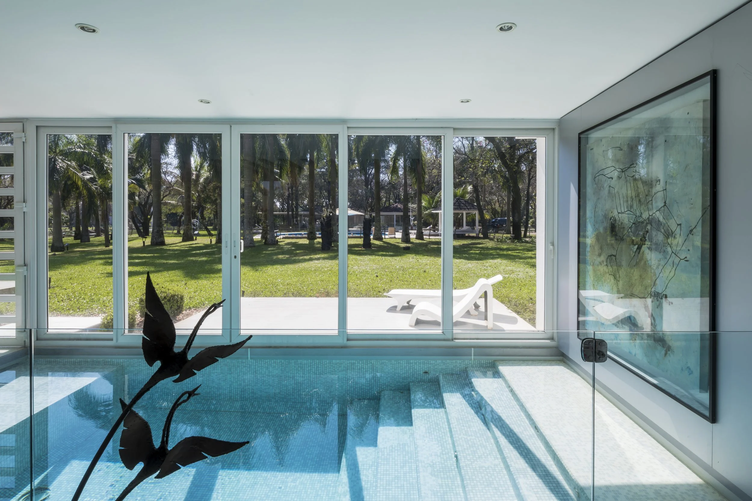 Vista de una piscina cubierta con vista a un jardín con árboles y sillas reclinables, a través de una gran ventana de vidrio